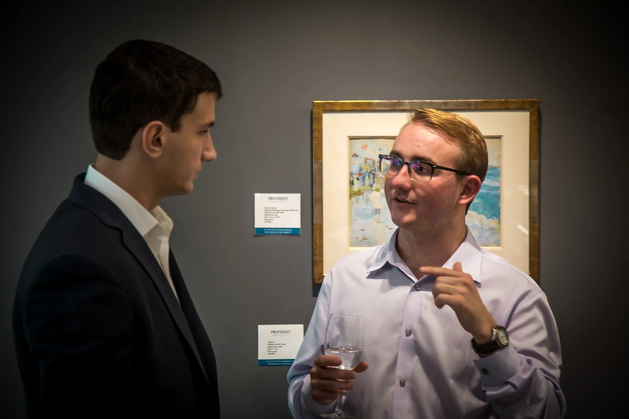 Two men are having a conversation at an art gallery or museum. One man is wearing a black suit, white shirt, with short dark hair, and is facing the other man. The other man, with glasses and short light brown hair, is holding a glass of water and ge