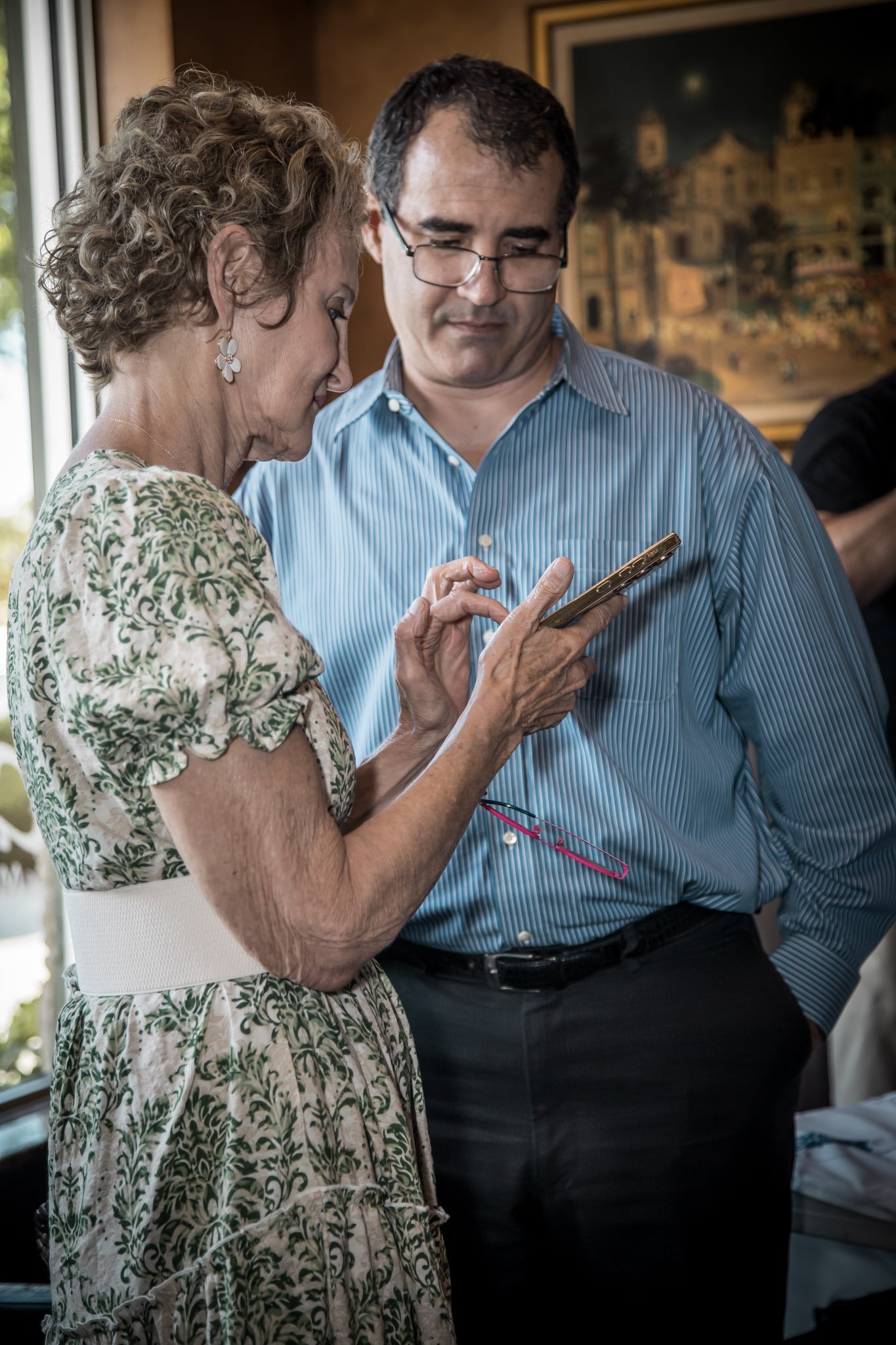 An elderly woman with curly hair and a floral dress shows something on her phone to a middle-aged man in glasses and a blue striped shirt, inside a warmly lit room with a painting in the background.