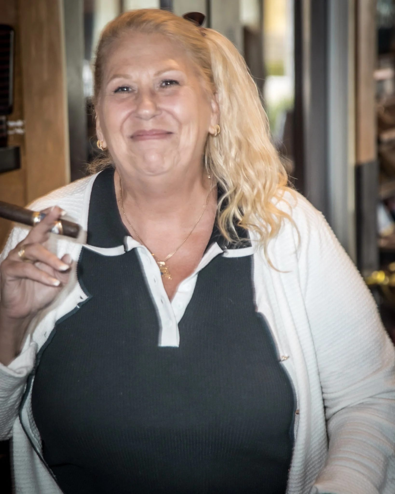 A smiling middle-aged woman with blonde curly hair holding a cigar near her face indoors, wearing a black and white top with jewelry.