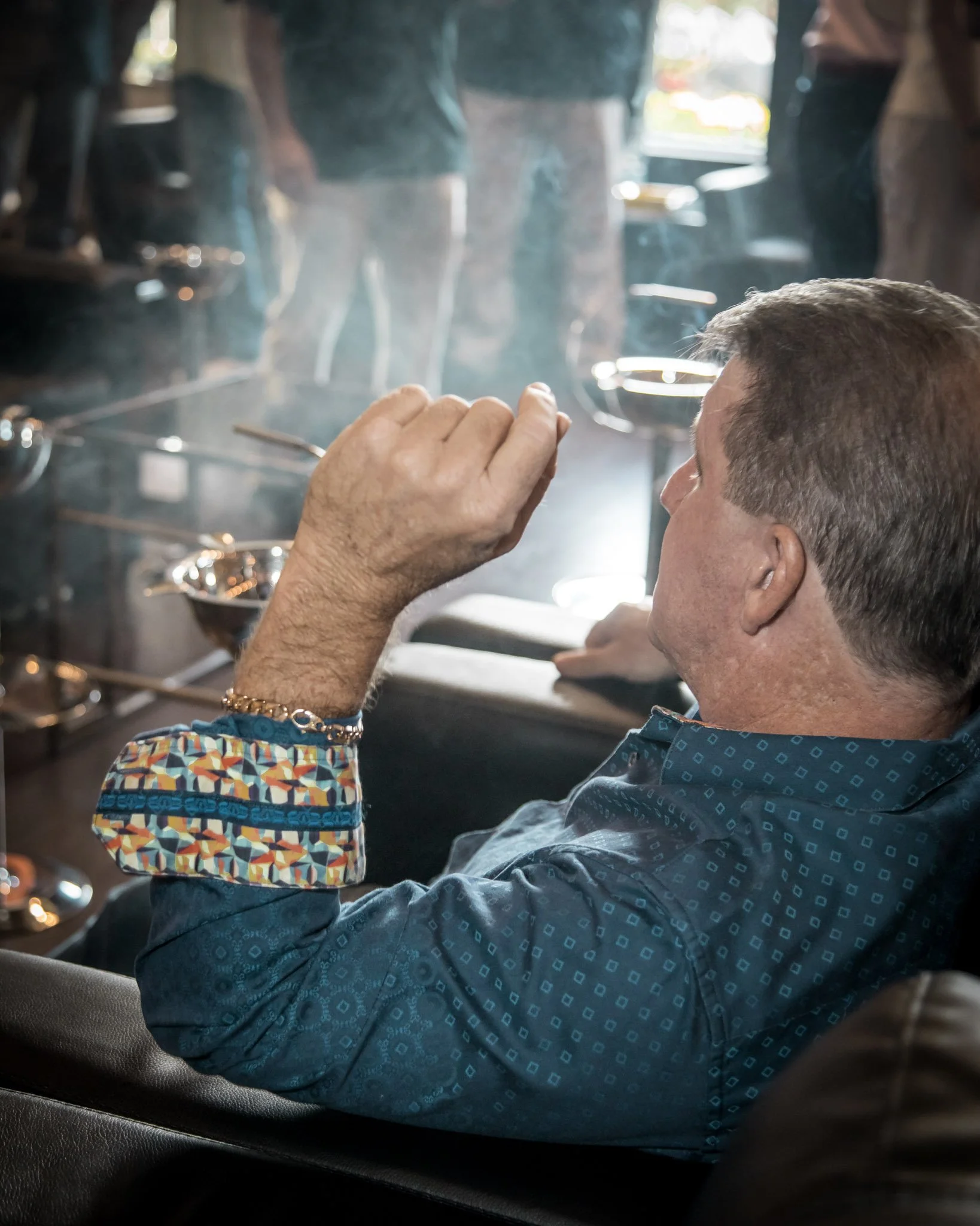 A man with short hair wearing a blue patterned shirt sitting on a sofa, holding a cigarette near his lips, with a smoky atmosphere in a room with other people in the background.