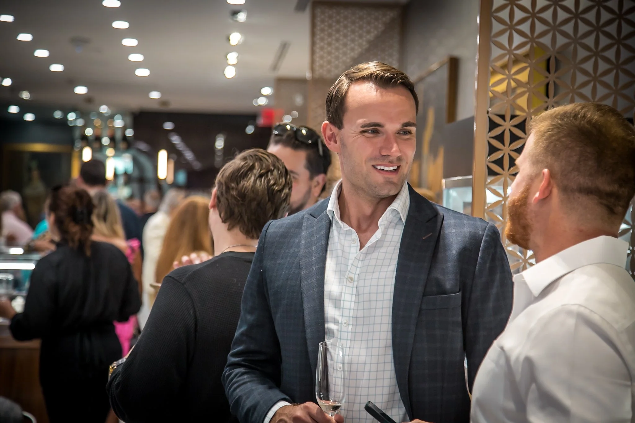 Two men engaged in conversation at a social gathering in a modern upscale setting, with a blurred background of other people and restaurant decor.