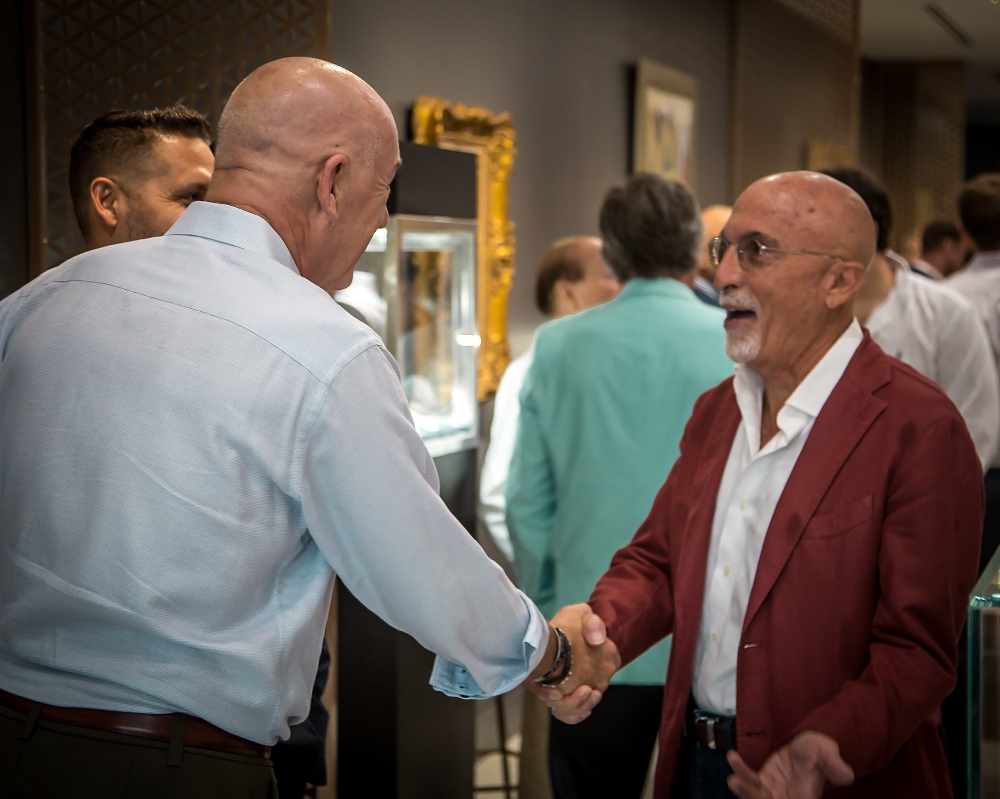 Two men shaking hands and smiling at each other at a social gathering, with other people in the background.