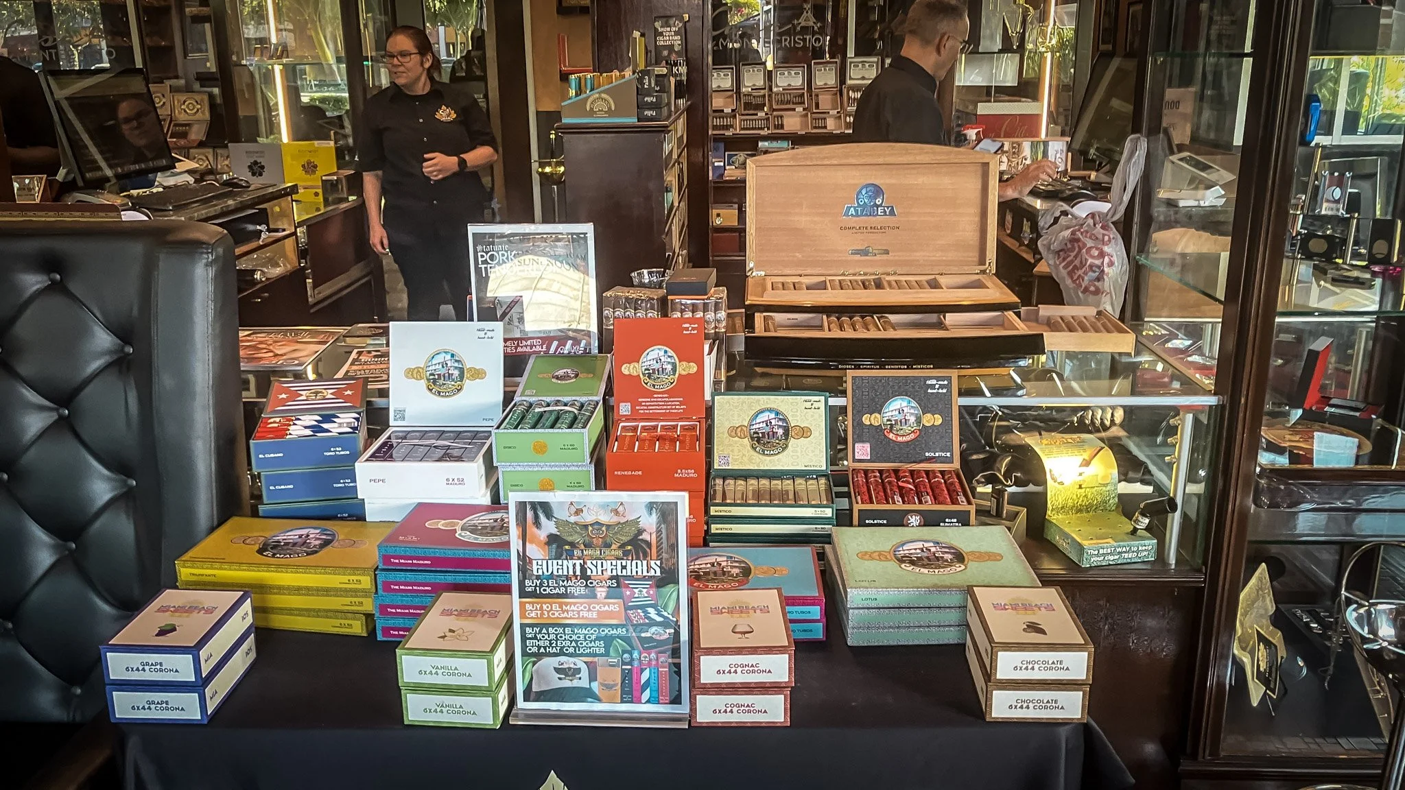 A display of various cigar boxes and packaging on a black table inside a store, with a glass display case and two employees working in the background.