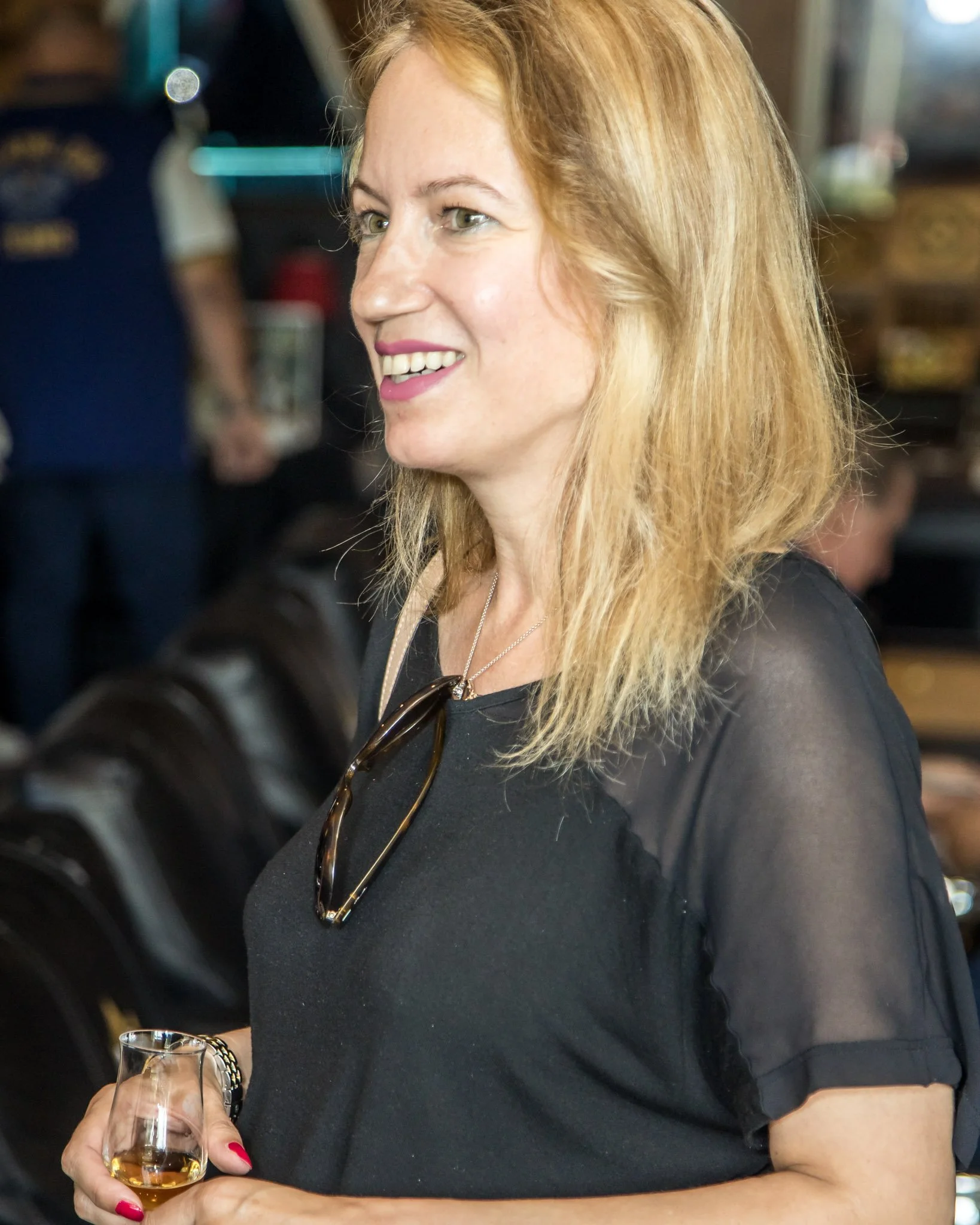 A woman with blonde hair smiling and holding a drink in a social setting, with blurred background showing other people and seating.