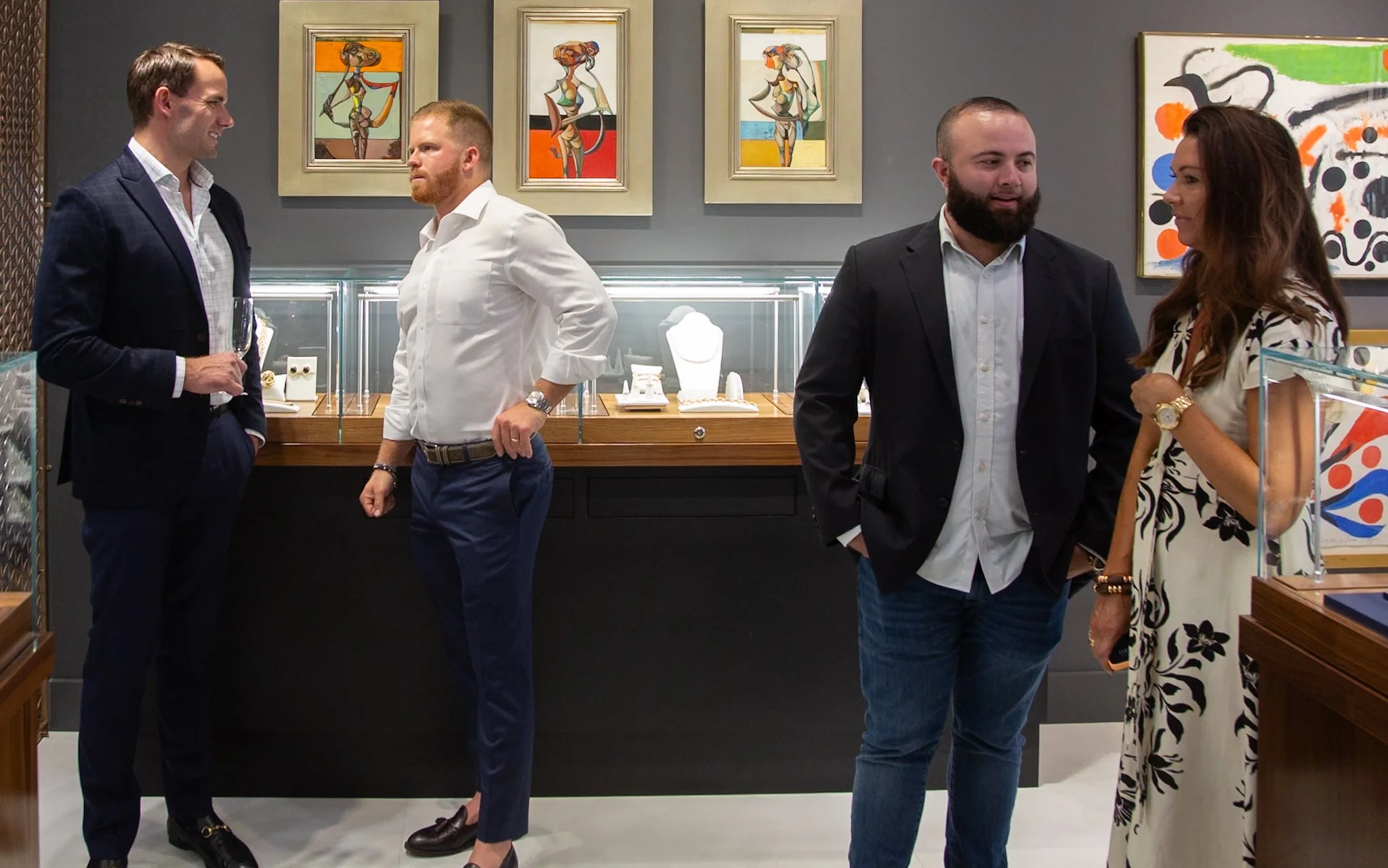 Four people are standing in an art gallery, two men with suits on the left and a man and woman conversing on the right. The background features colorful abstract and figurative artwork.
