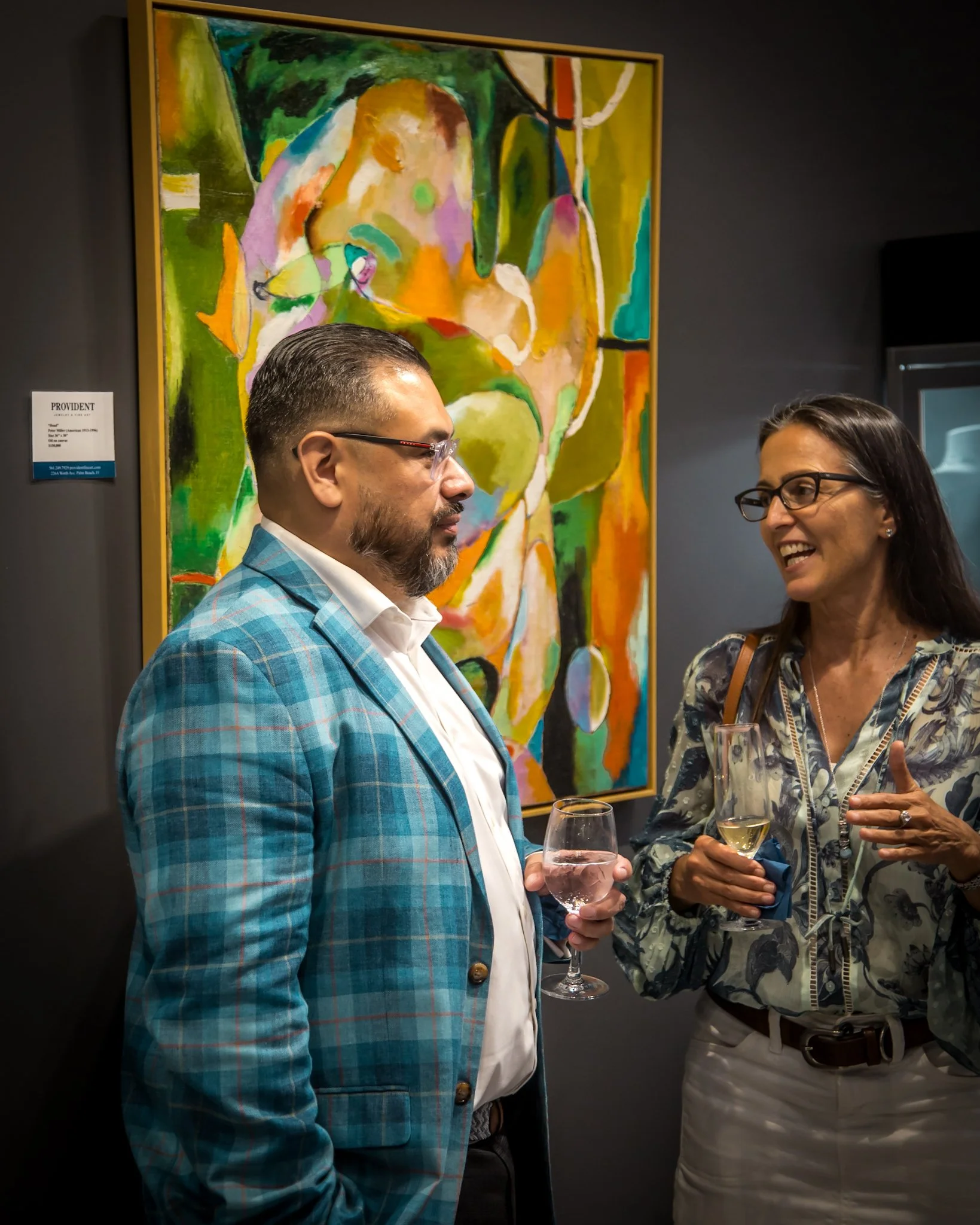 Two people, a man in a blue plaid blazer and a woman in a floral blouse, are having a conversation at an art gallery, holding glasses of wine. A colorful abstract painting is in the background.