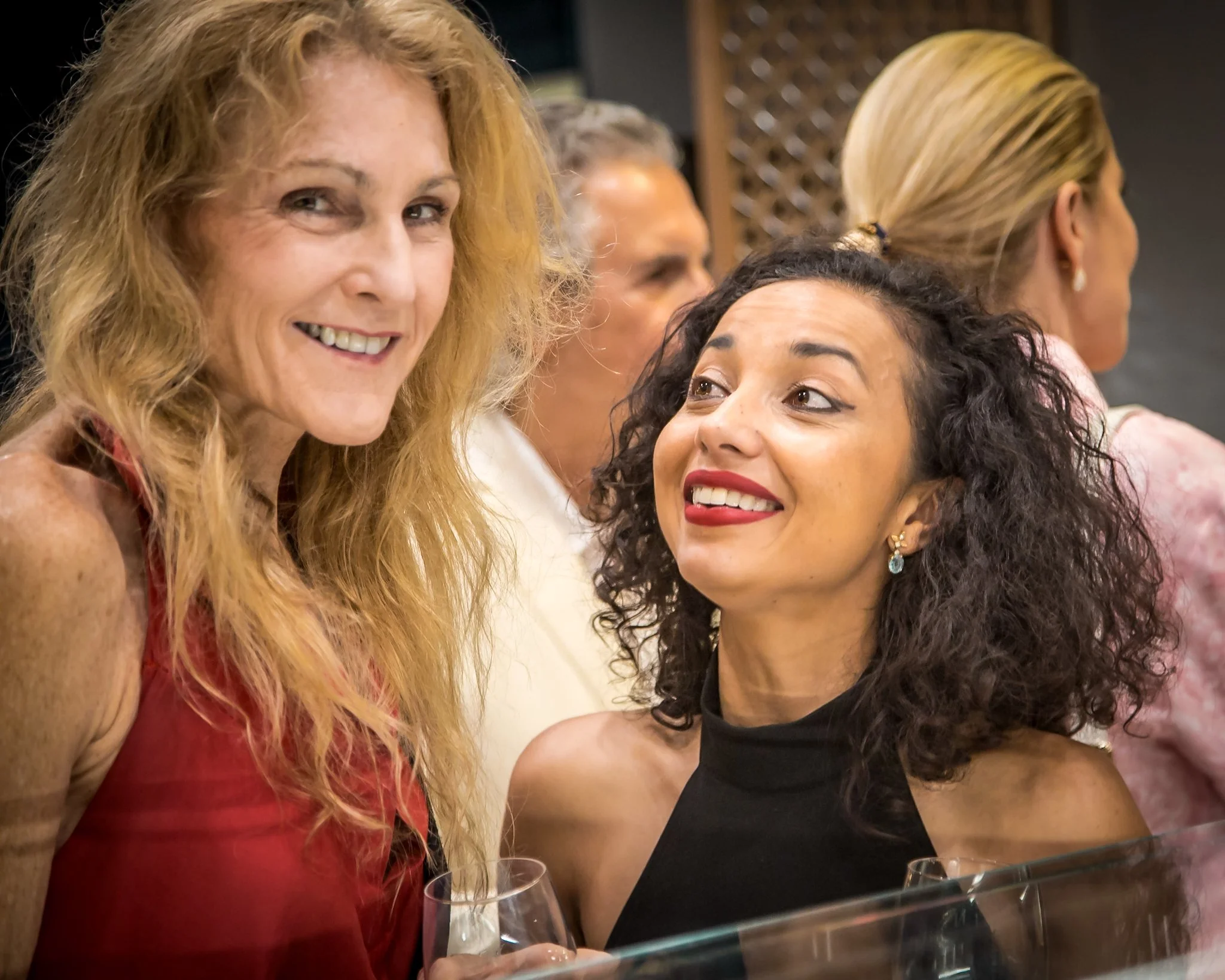 Two women smiling and talking at an indoor social event, with others in the background.