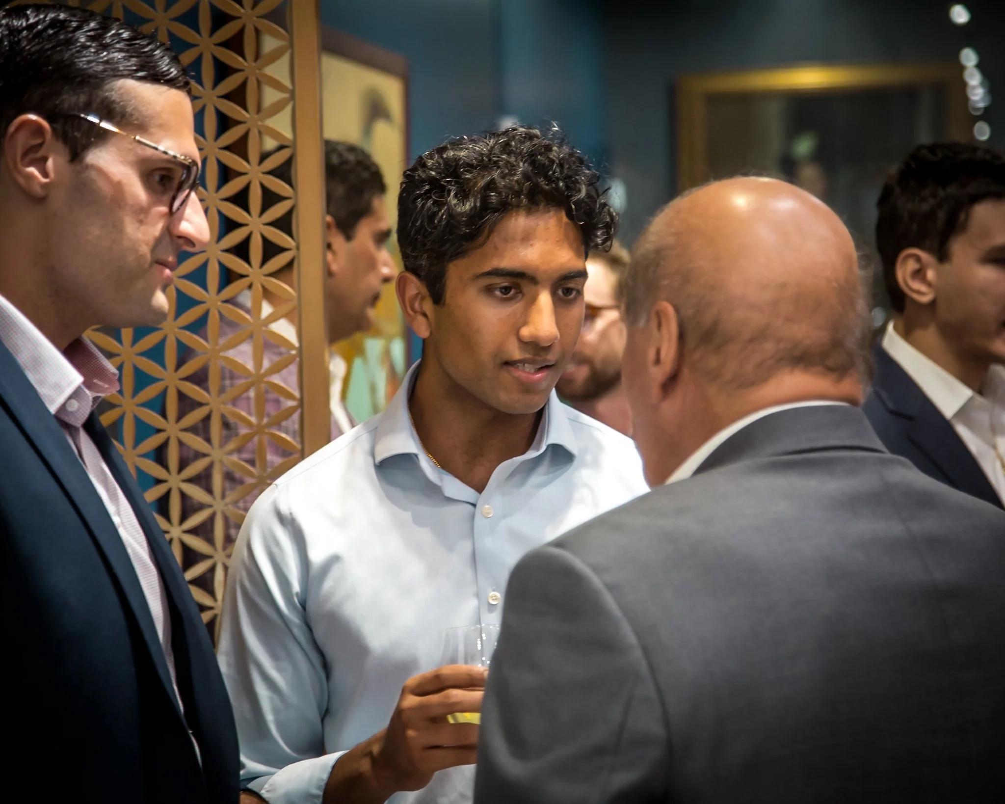 Group of men in business attire socializing at a formal event in a room with decorative wall panels and framed pictures.