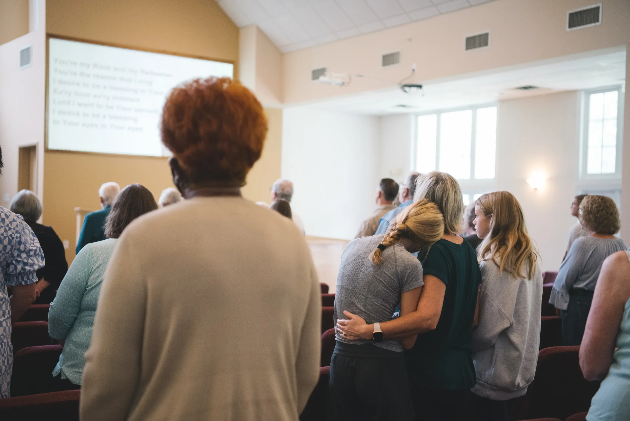 People attending a church service, with some sitting and others standing, and a large screen displaying lyrics in the background.