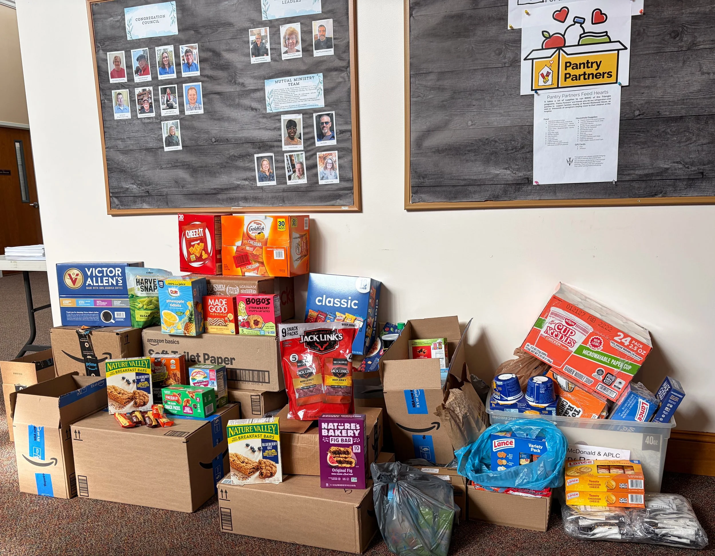 Table filled with boxes and packages of food and supplies, with a bulletin board in the background displaying photographs and informational posters related to community services and leadership.