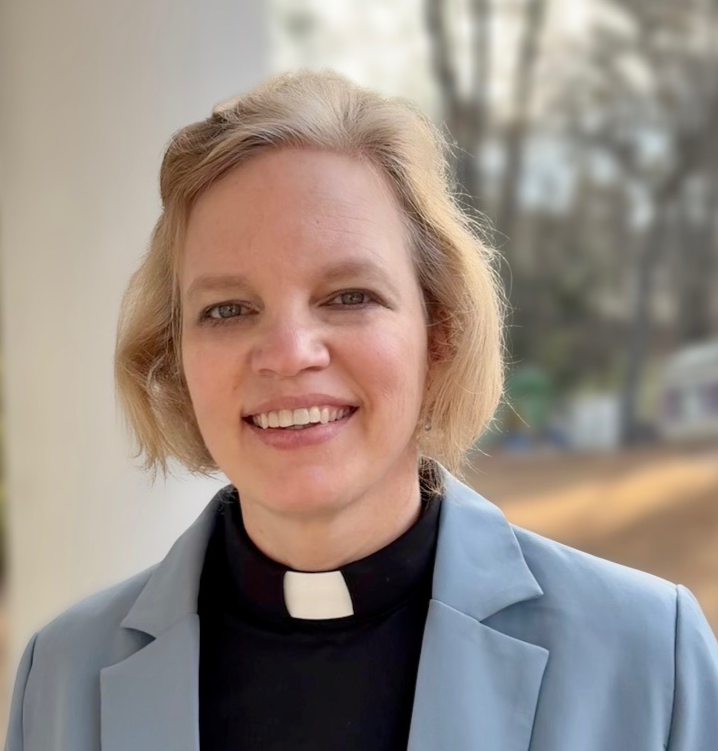 Portrait of a smiling female priest with short blonde hair, wearing a black clergy collar and a light blue blazer, standing outdoors with trees and a residential area in the background.