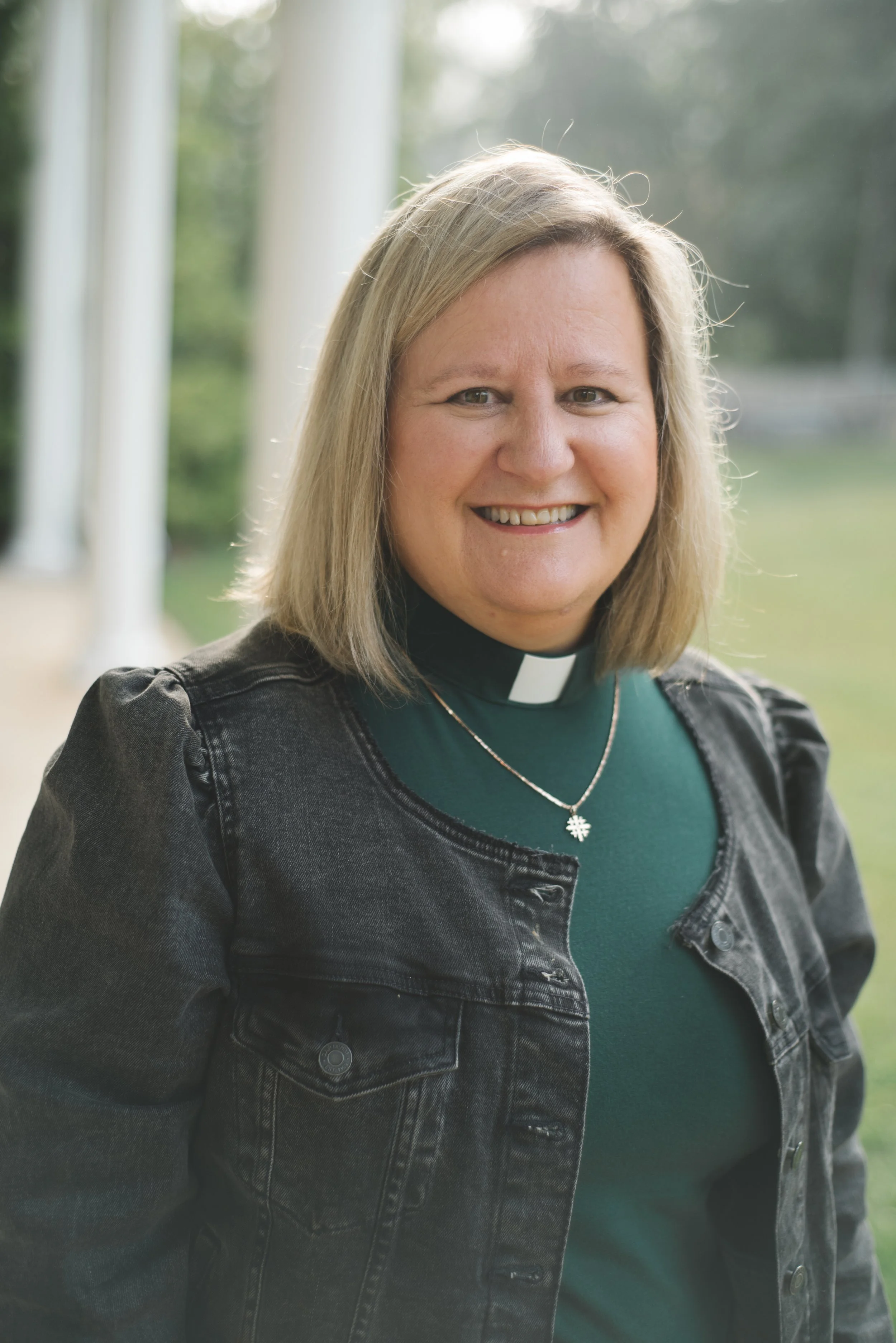 A woman wearing a clerical collar and a black denim jacket, smiling outdoors with trees and a porch in the background.