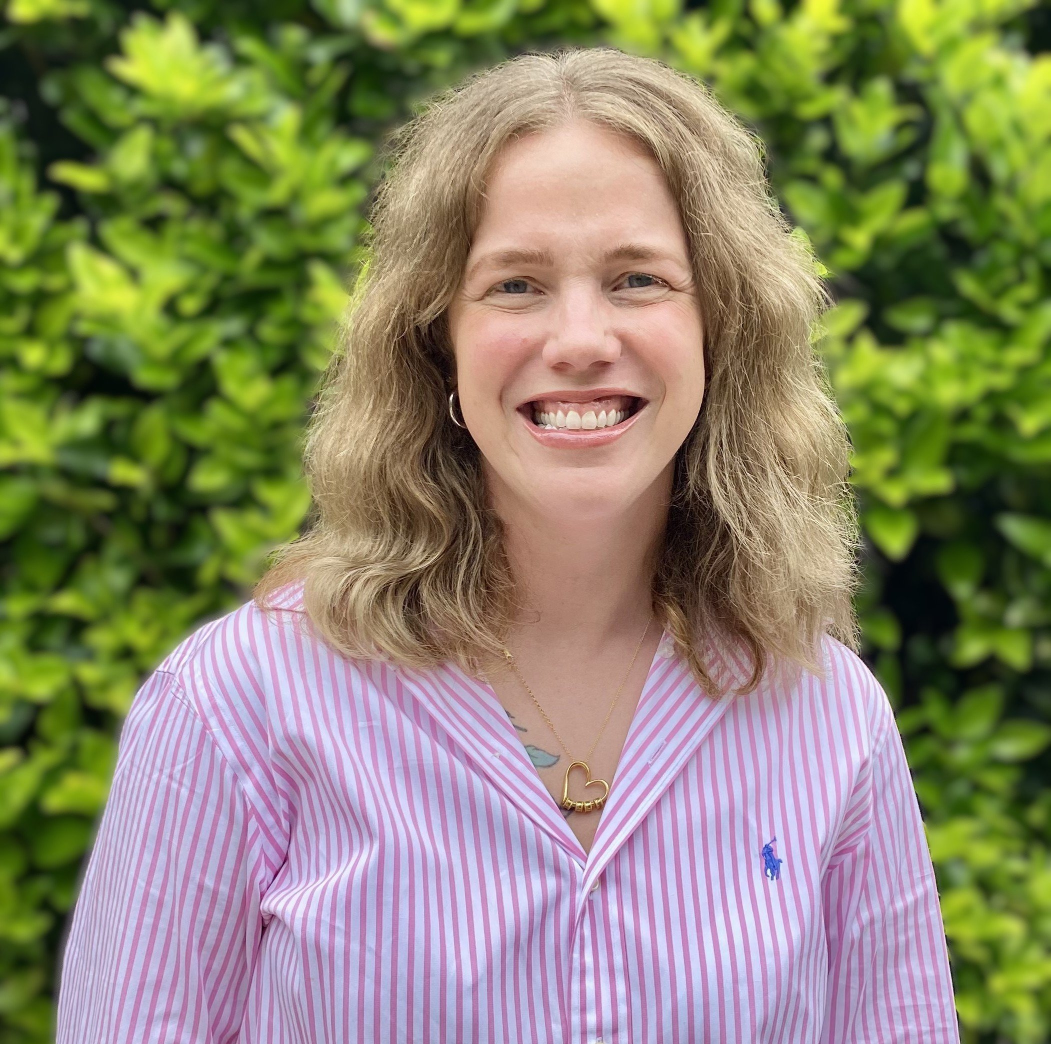 A woman with shoulder-length curly blonde hair smiling, wearing a pink and white striped shirt with a small blue logo, a gold heart-shaped necklace, and silver hoop earrings, standing outdoors with green foliage background.