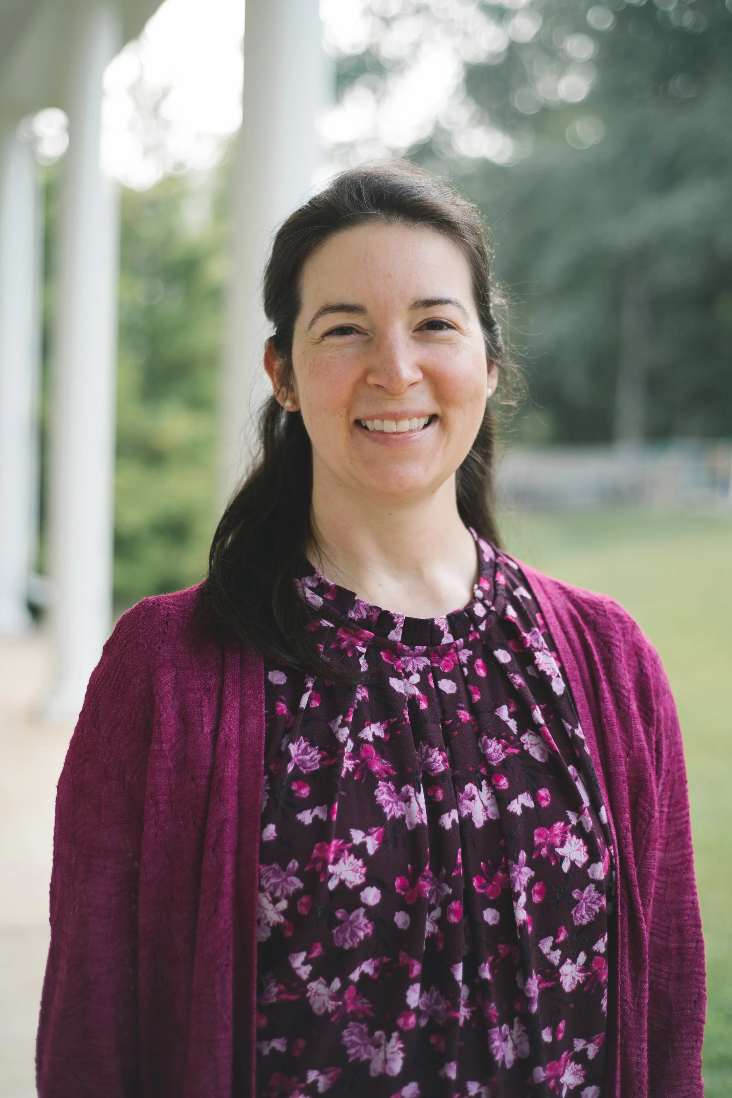 A woman with dark brown hair smiling outdoors, wearing a purple cardigan and a black shirt with pink and purple floral patterns.