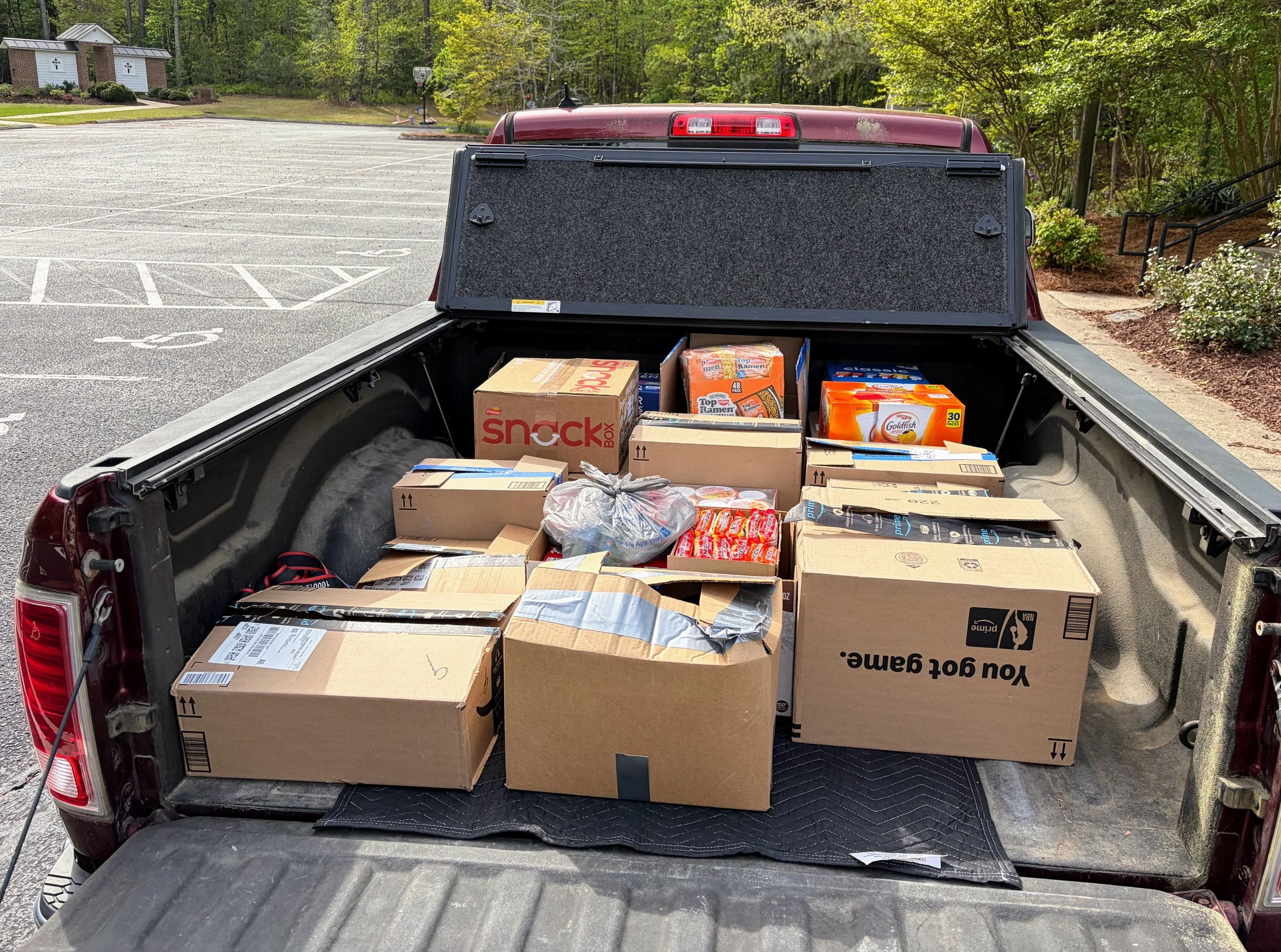 Pickup truck bed filled with boxes, some labeled as snacks and instant ramen, with a suburban background of a parking lot, green trees, and a small building.