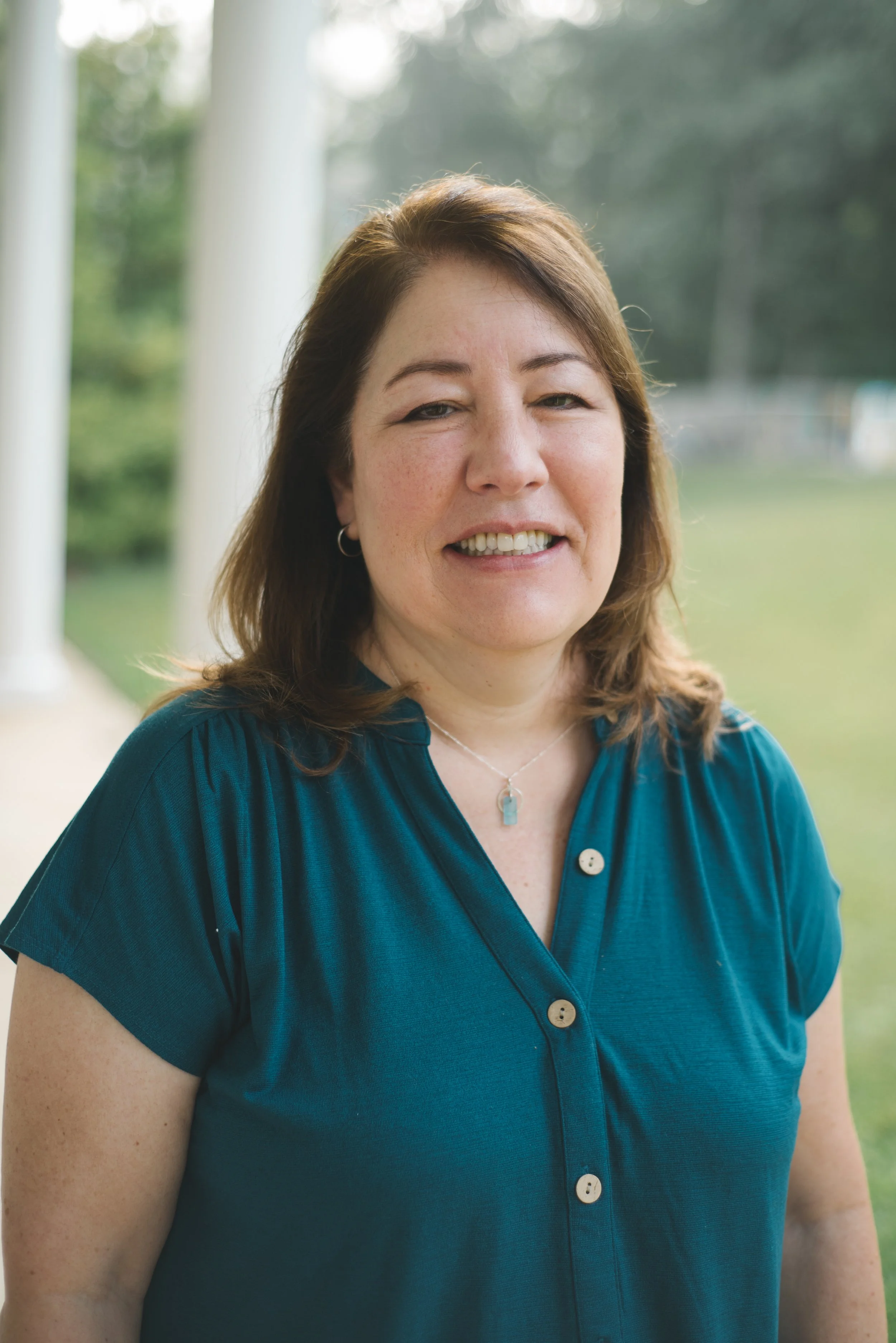 A woman with shoulder-length brown hair smiling outdoors, wearing a teal blouse with buttons and a silver necklace, standing on a porch with white columns and greenery in the background.