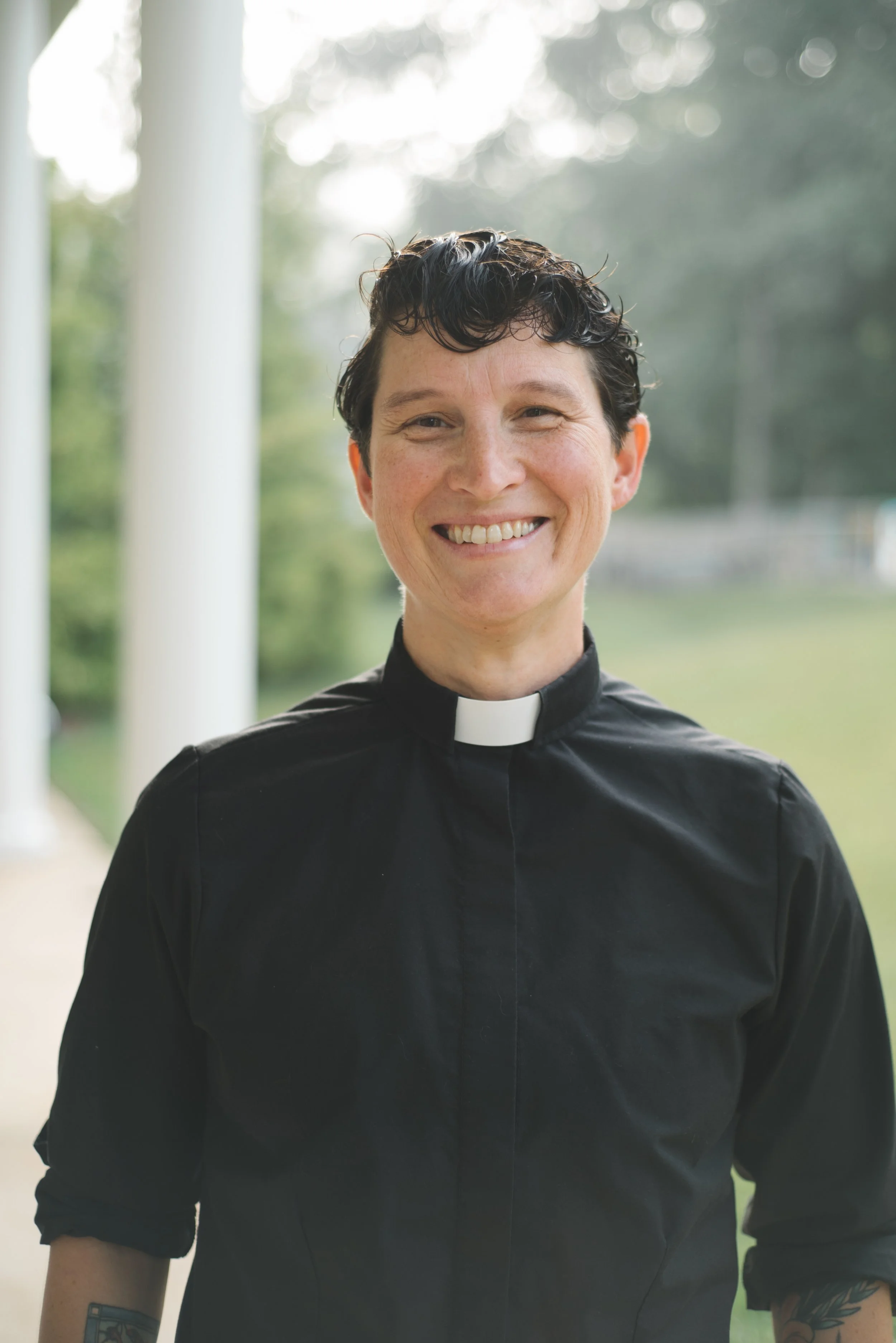 A smiling woman dressed in a black clerical shirt with a white collar, standing outdoors on a porch with a blurred background of trees and sky.