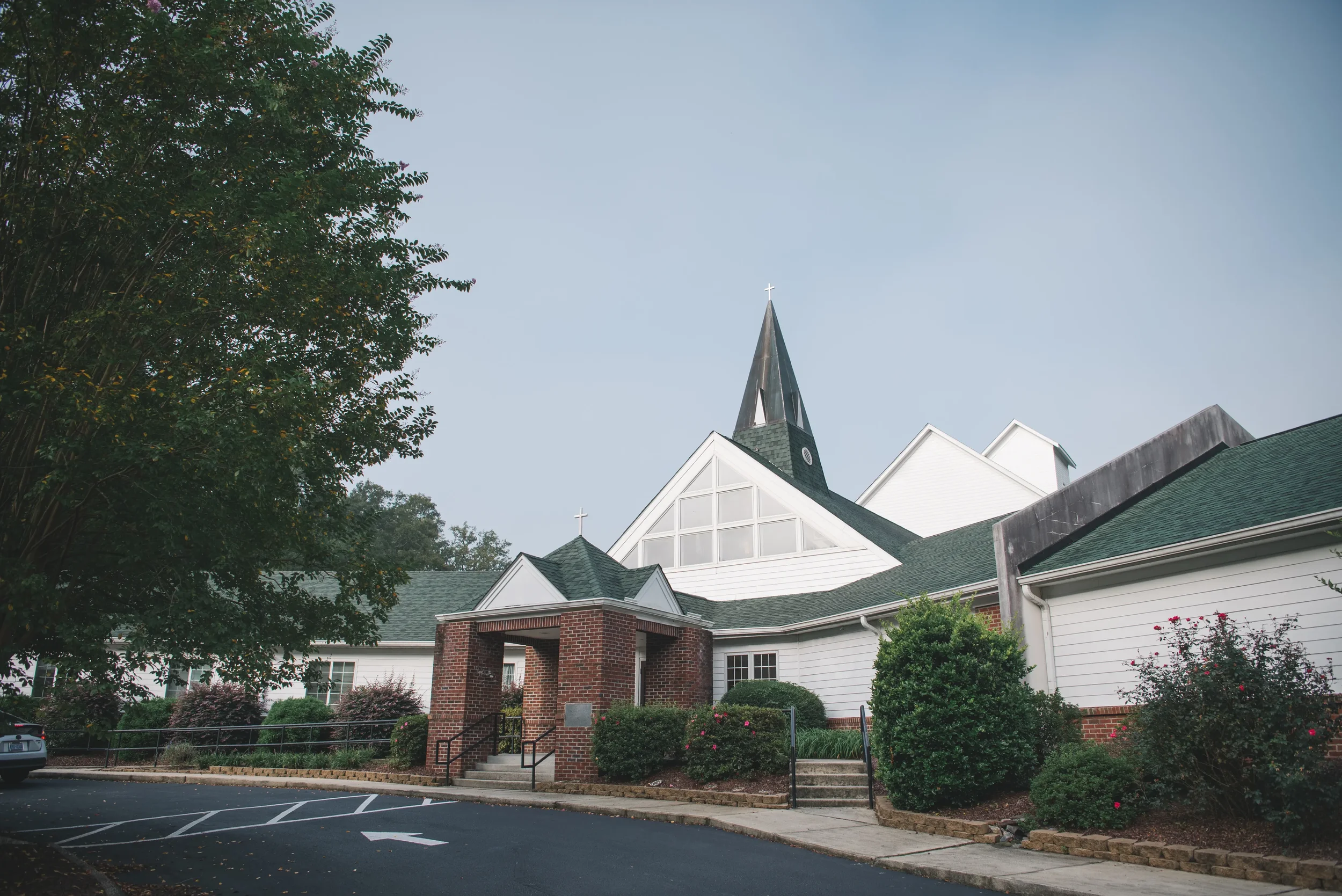 APLC church with white exterior walls, green roofing, and a tall steeple with a cross on top. The church has decorated bushes and trees in front, along with a small brick entrance and a parking lot nearby.