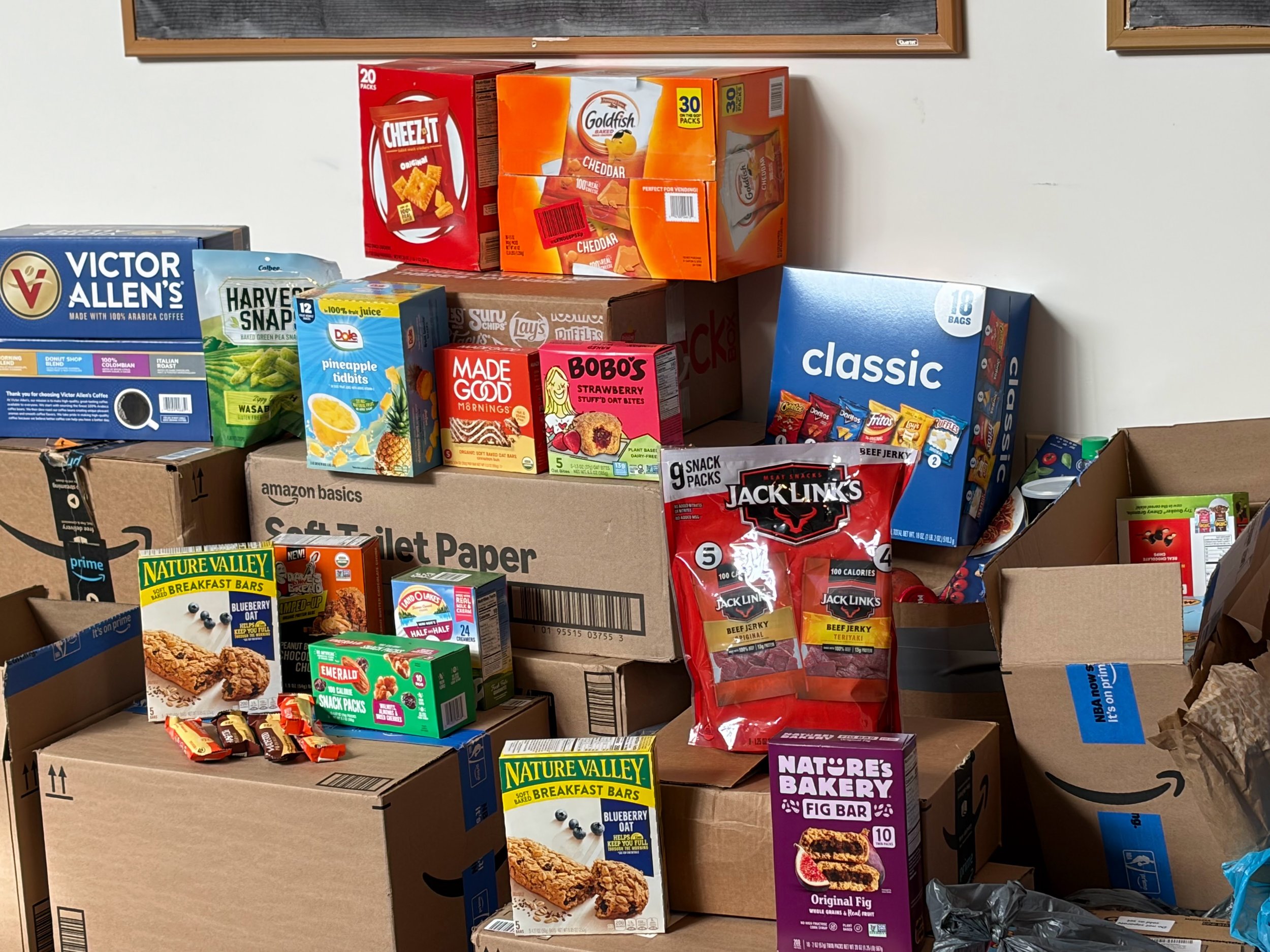 Various snack boxes and food items stacked on top of cardboard boxes, including Cheez-It, Goldfish, Nature Valley breakfast bars, Jack Link's beef jerky, and other snack packages.