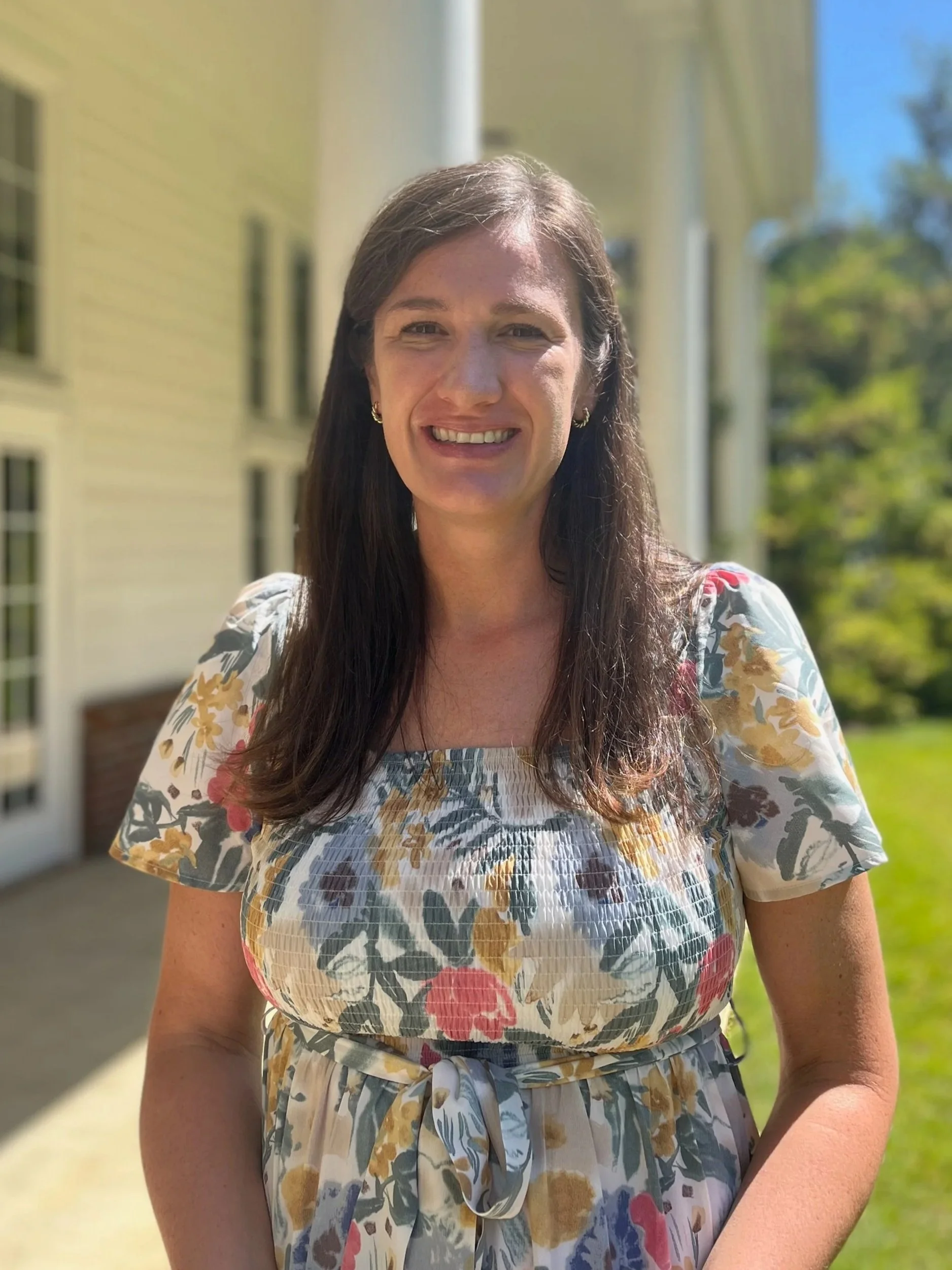 A woman with long dark hair smiling outdoors in front of a yellow house with white trim and a green lawn.