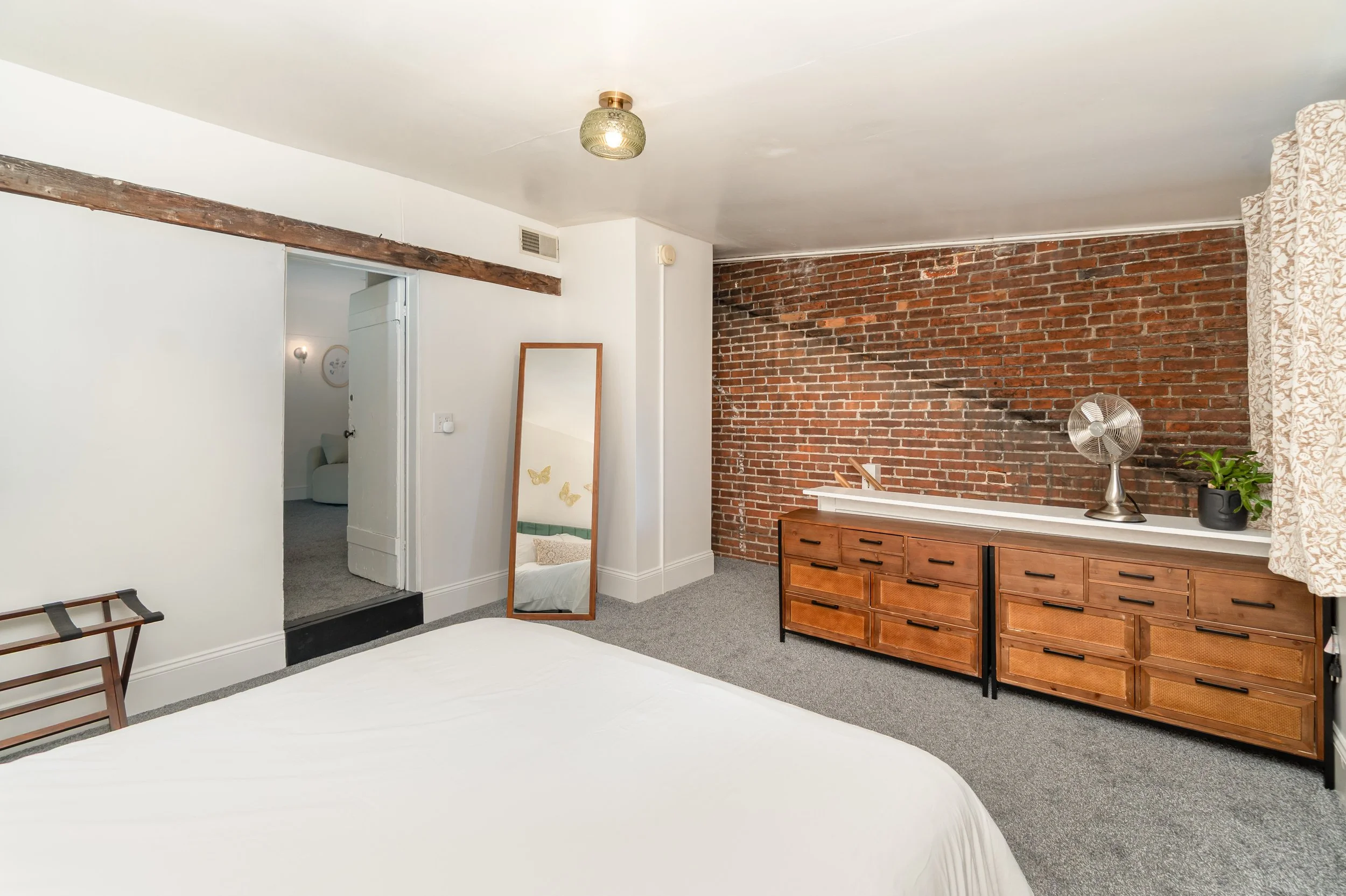 Bedroom with white walls, a large bed with white bedding, a wooden dresser, a mirror, a small fan, and a brick accent wall with a plant and curtain.