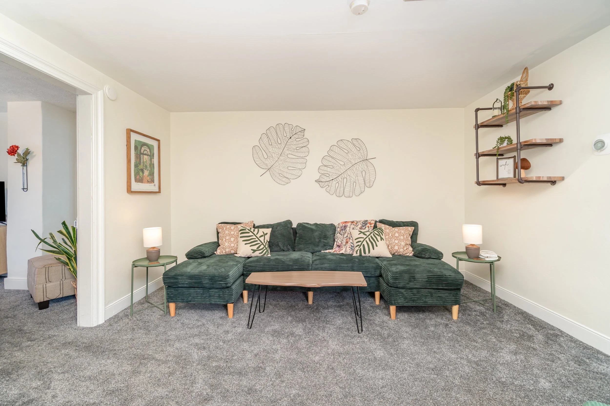 Living room featuring a green velvet sectional sofa with patterned and leaf-themed pillows, a wooden coffee table, two side tables with lamps, framed artwork, wall-mounted decorative shelves with plants, and wall art of wire leaf outlines.