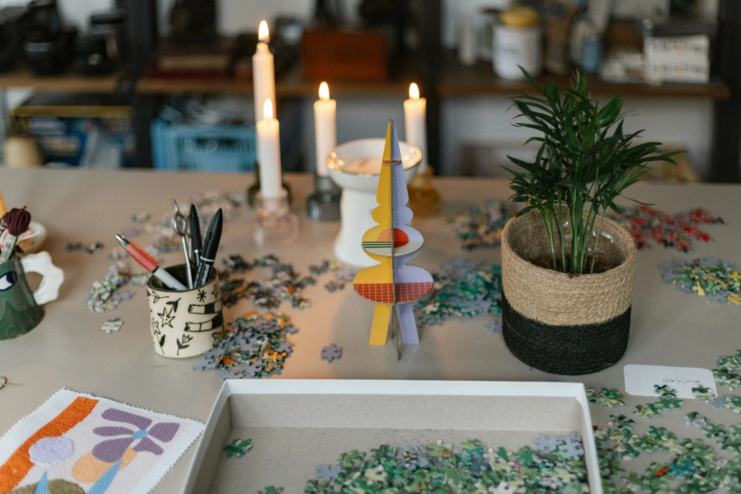A cluttered table with scattered puzzle pieces, a partially completed puzzle, pens, and a plant in a decorative pot. There are candles and a colorful paper decoration in the background.