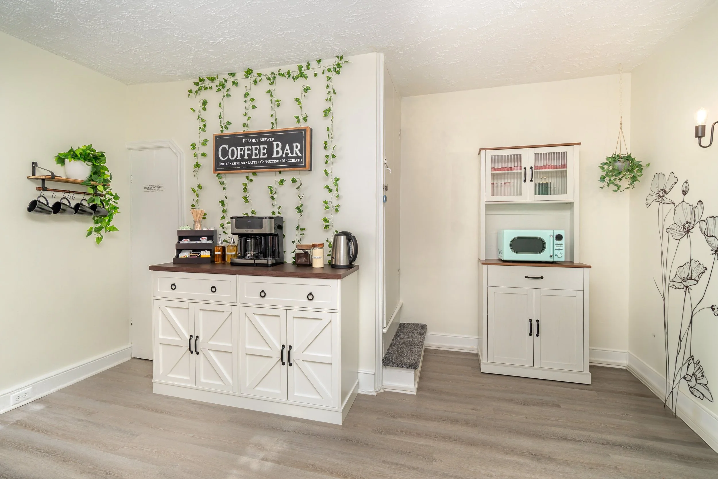 A coffee station in a cozy kitchen with white cabinets, a coffee maker, teapot, and jars on the counter, green hanging plants, black cups on a wall shelf, and a mint microwave on a white cabinet, with wall art and lighting.