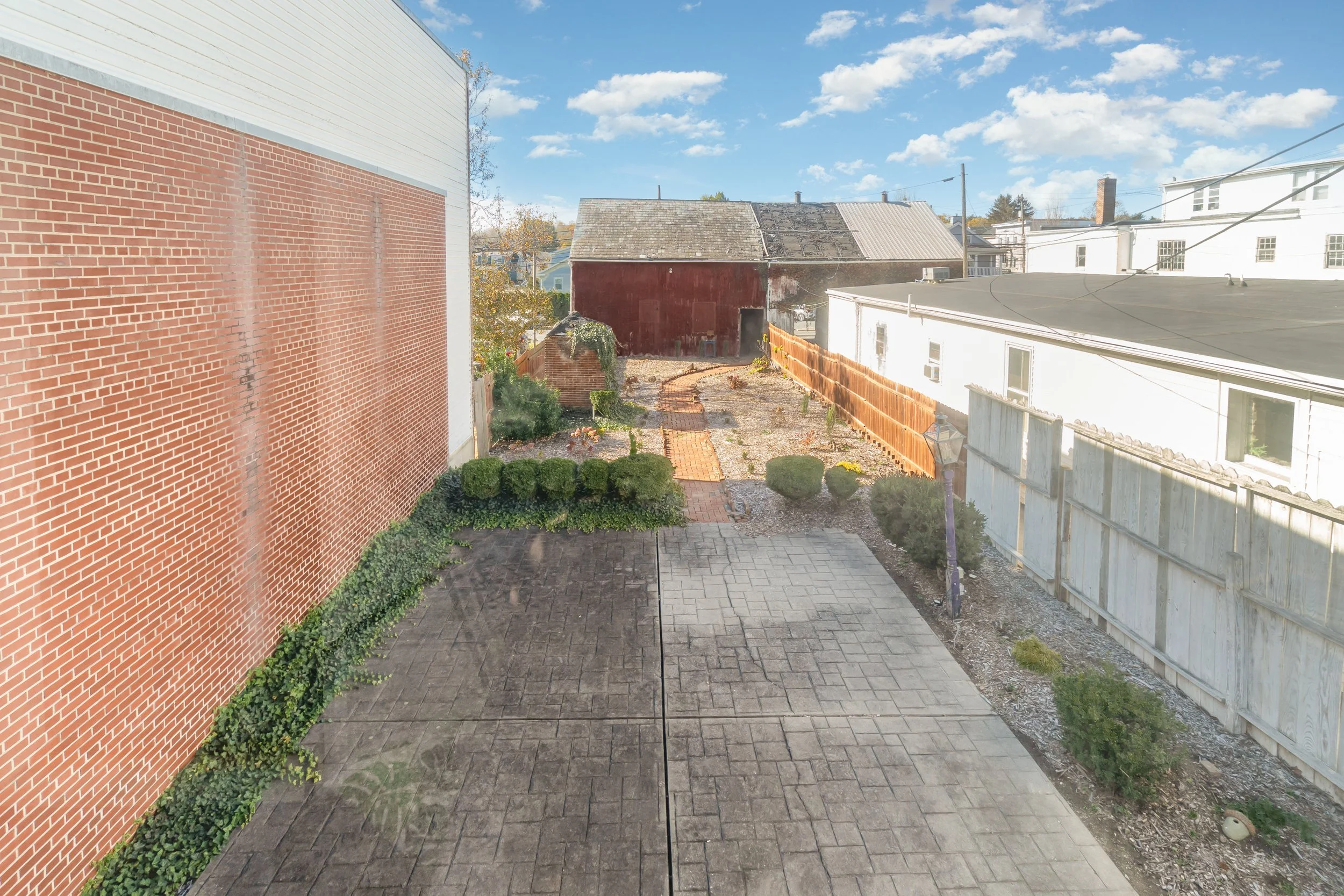 View of a backyard with a paved patio, small shrubs, and plants along a fence and house wall, with an old red barn at the end of the yard under a partly cloudy sky.