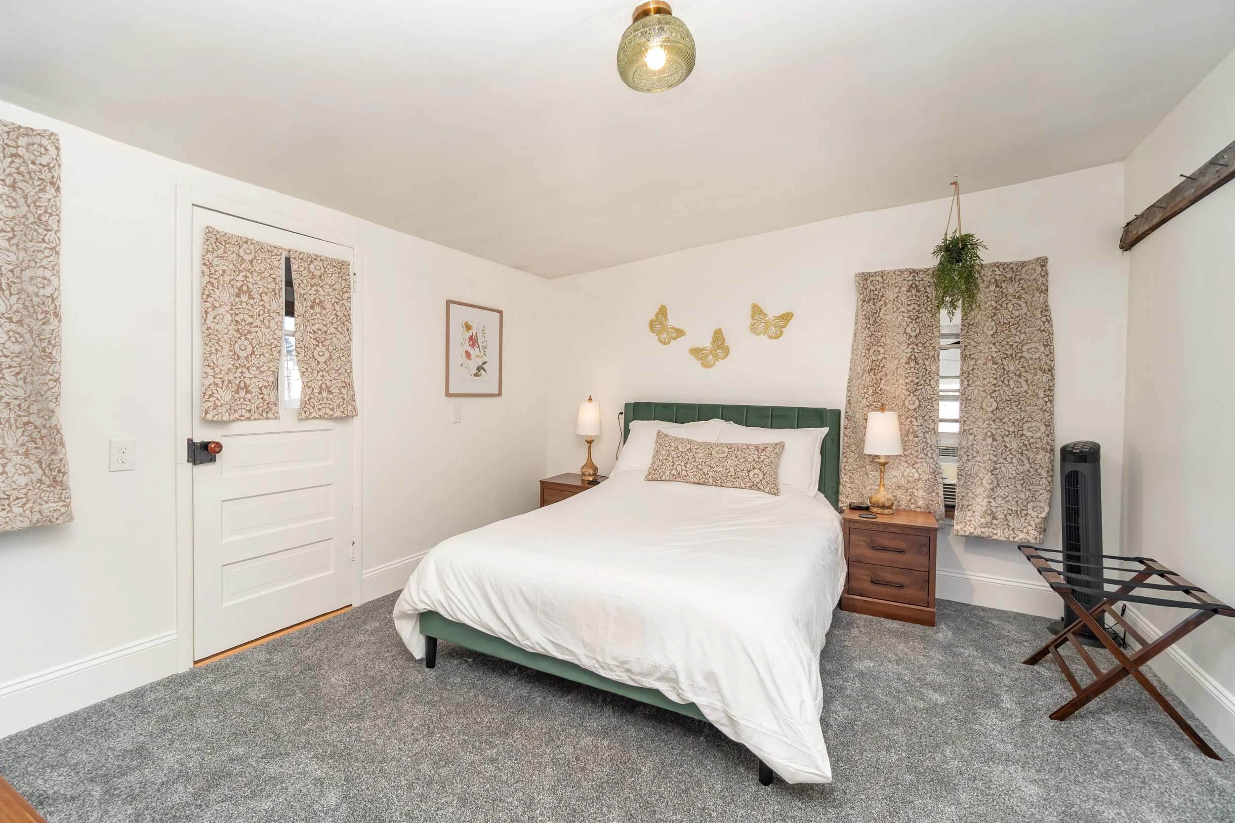 Bedroom with a green headboard, white bedding, two wooden nightstands with lamps, beige patterned curtains, a plant hanging from the ceiling, and framed artwork with butterflies on the wall.