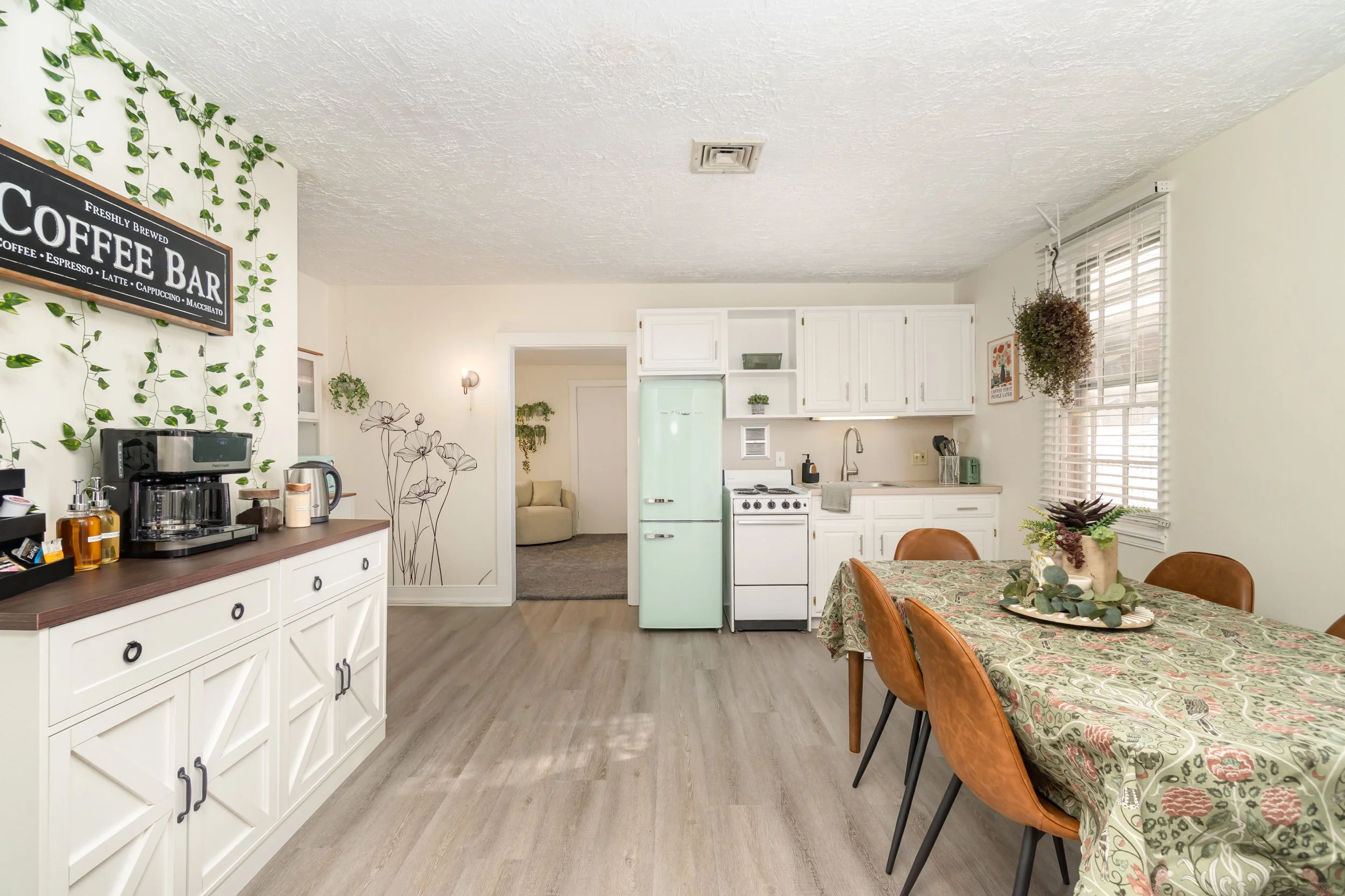 A kitchen and dining area with white cabinets, a light green retro refrigerator, a table with a floral tablecloth, and brown chairs. The wall facing the table has a hanging potted plant and a window with blinds. There is a sideboard with a coffee maker, kettle, and bottles, and a wall sign that reads "Coffee Bar." In the background, a doorway leads to a seating area with a beige chair and wall art.