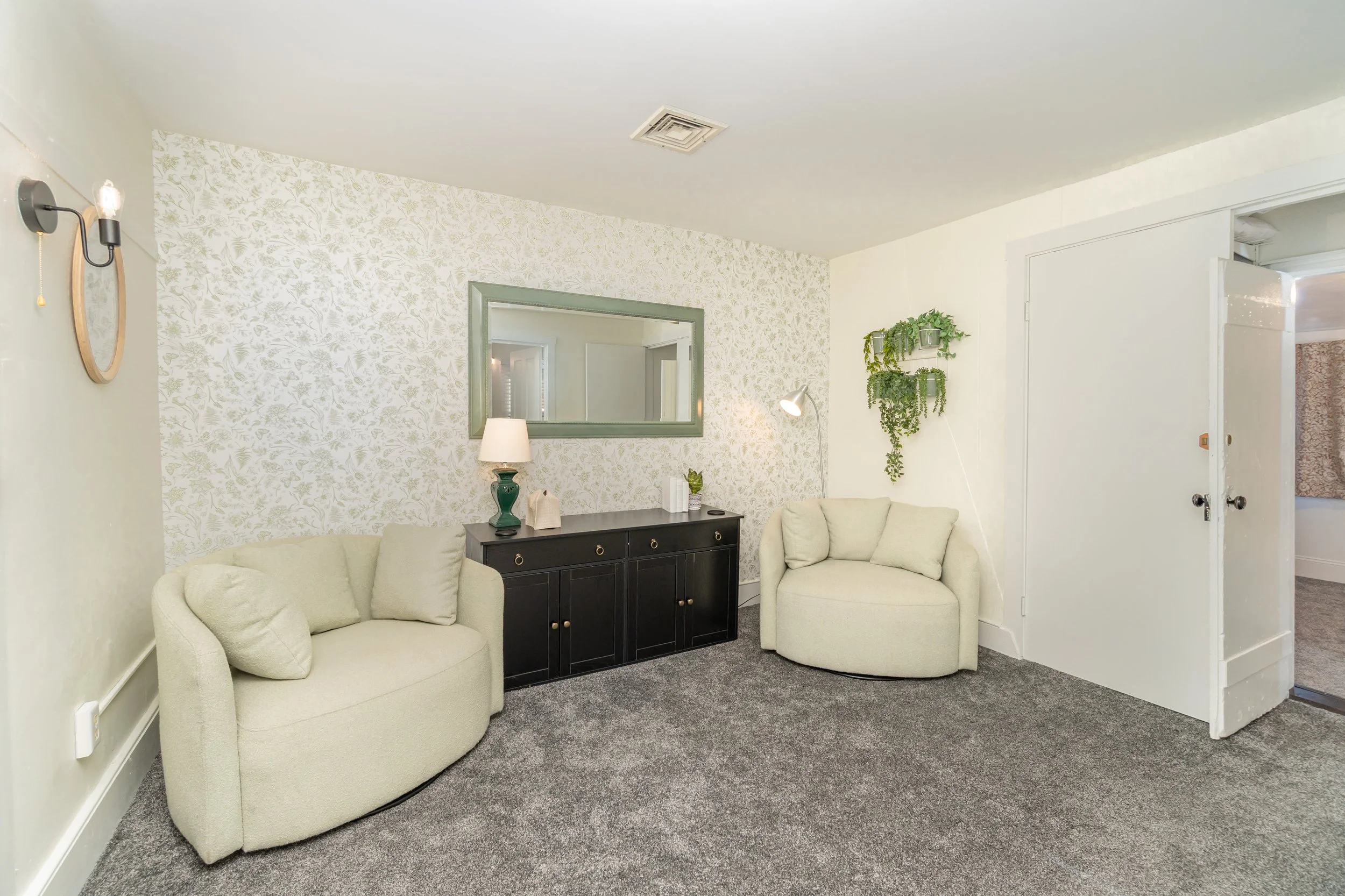 Living room with two cream-colored upholstered couches, a black sideboard with a mirror above, a green table lamp, and a hanging plant on the wall.
