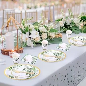 Elegant outdoor table setting with white and green floral centerpiece, gold-rimmed plates with utensils, white candles in glass holders, and decorative lanterns on a white tablecloth.