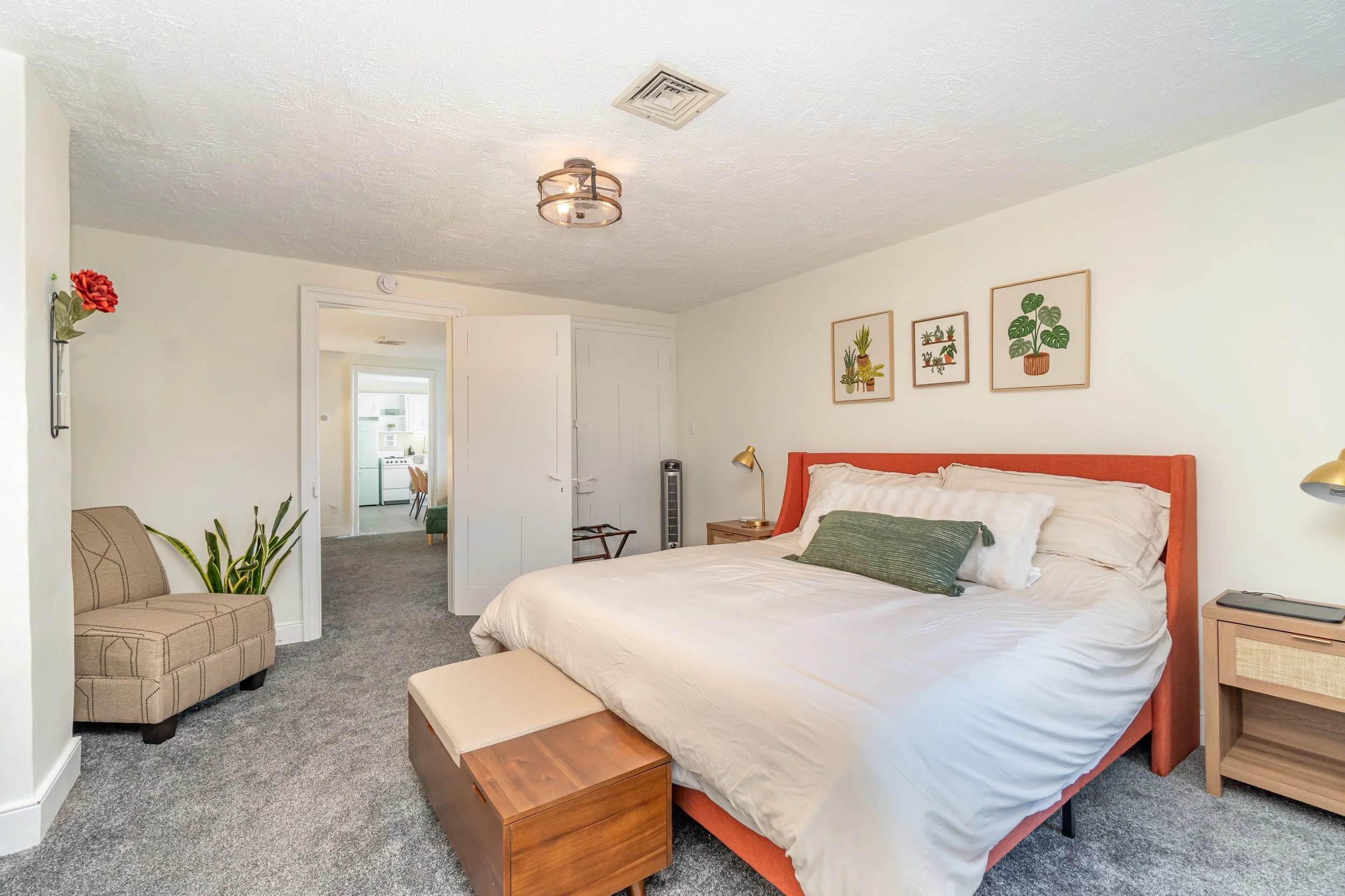 Bedroom with a large bed, orange headboard, wooden nightstands, framed leaf art on wall, green pillow, beige walls, gray carpet, small bench at foot of bed, small sofa, plants, and door leading to another room.