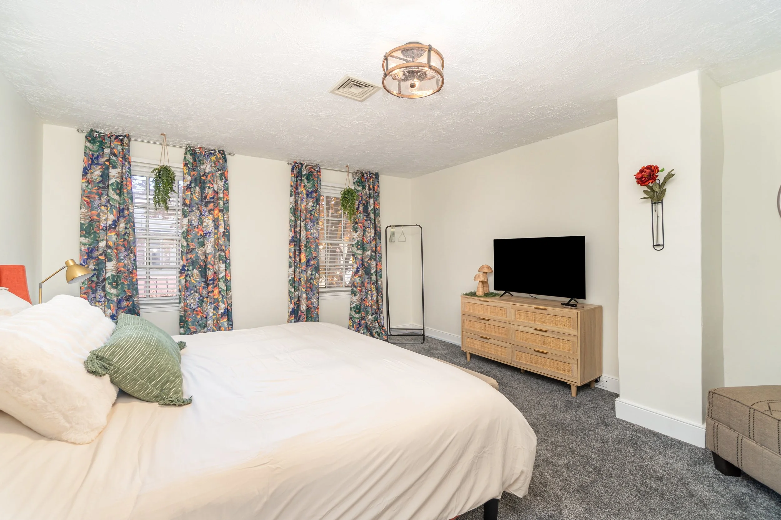 A bedroom with a large bed, green pillow, floral curtains on two windows, a flat-screen TV on a rattan dresser, a floor lamp, and wall decor including a red flower in a black vase.