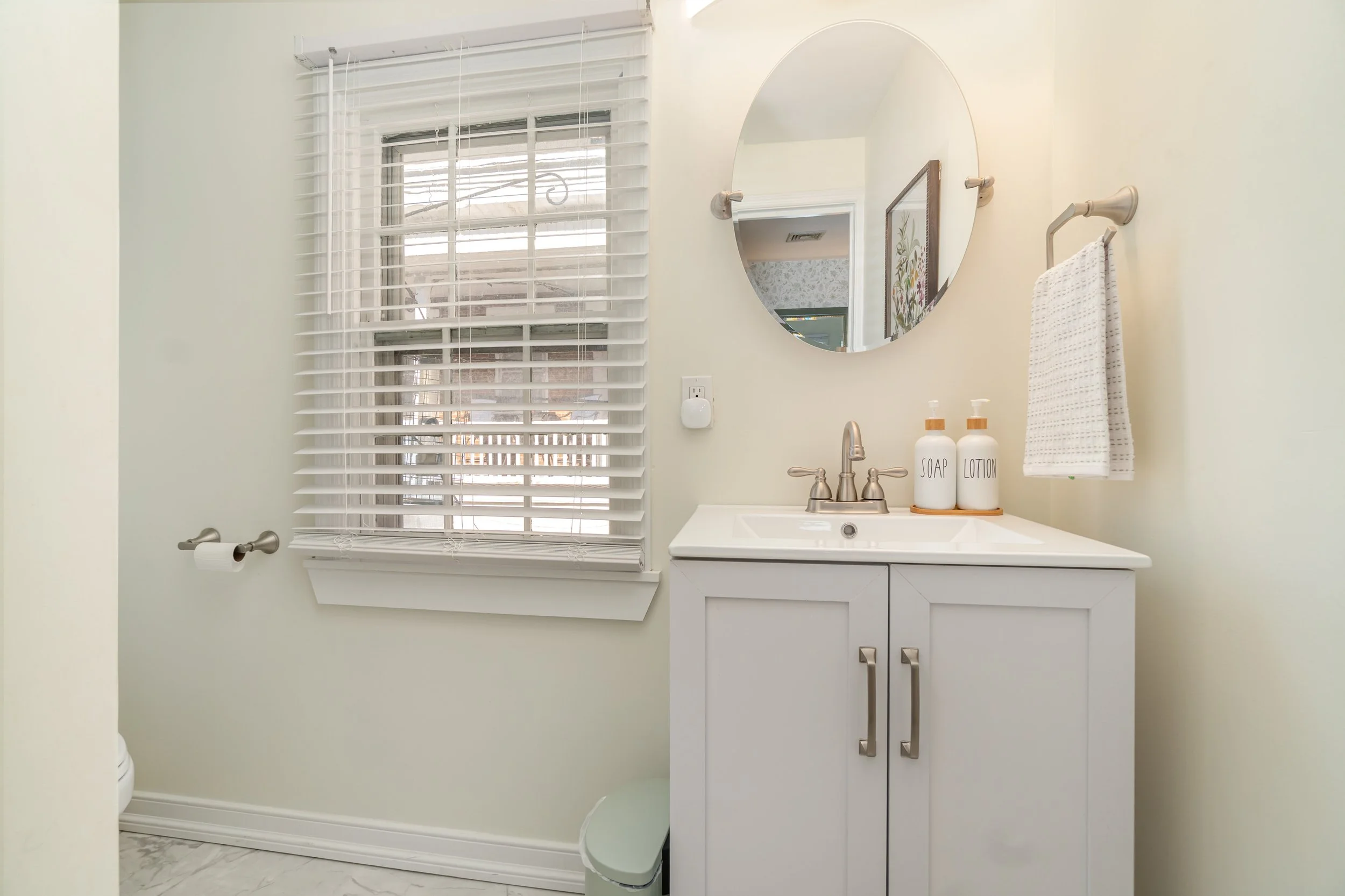 A small bathroom with a white vanity, oval mirror, and light-colored walls. There are soap and lotion dispensers on the sink, a towel on a wall-mounted rack, and a window with white blinds.