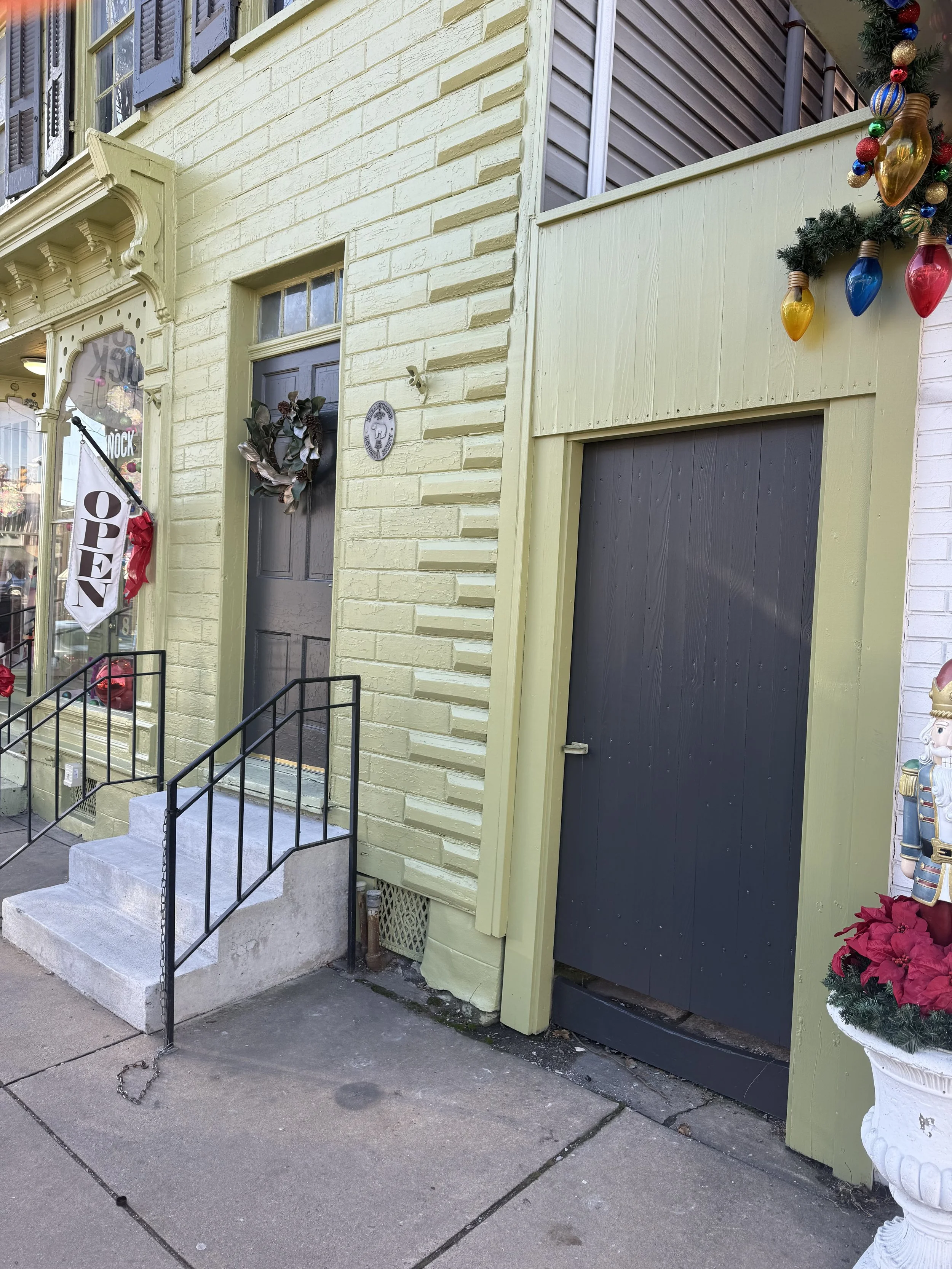 A storefront with a yellow painted brick exterior, decorated with holiday ornaments, a wreath on the door, and a sign that reads 'OPEN'. There are concrete steps with black metal railings leading up to the door, and a Christmas nutcracker figure with