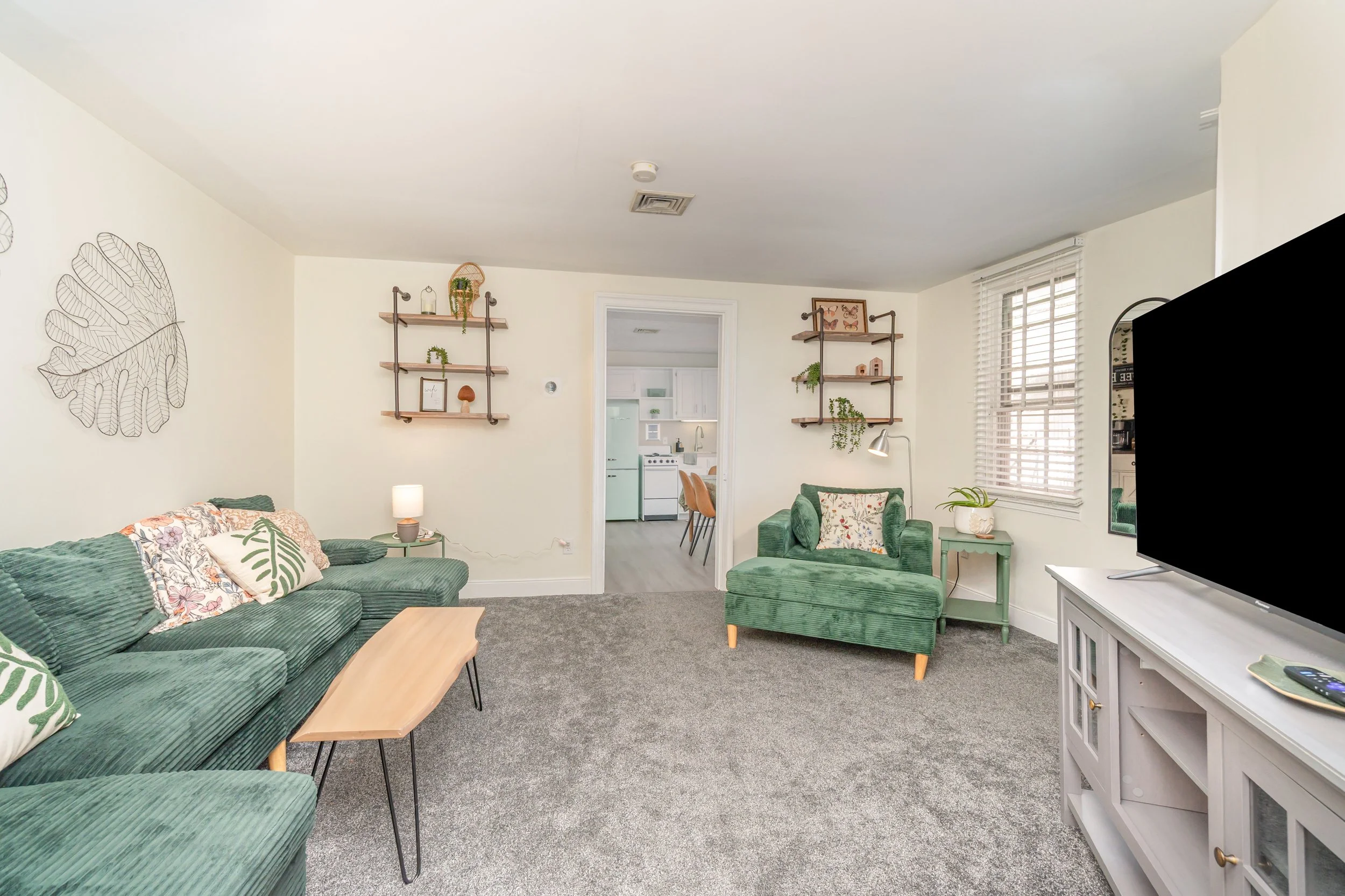 Living room with green sofa, green armchair, white TV stand, wall shelves, and décor, with an open door leading to a kitchen visible in the background.