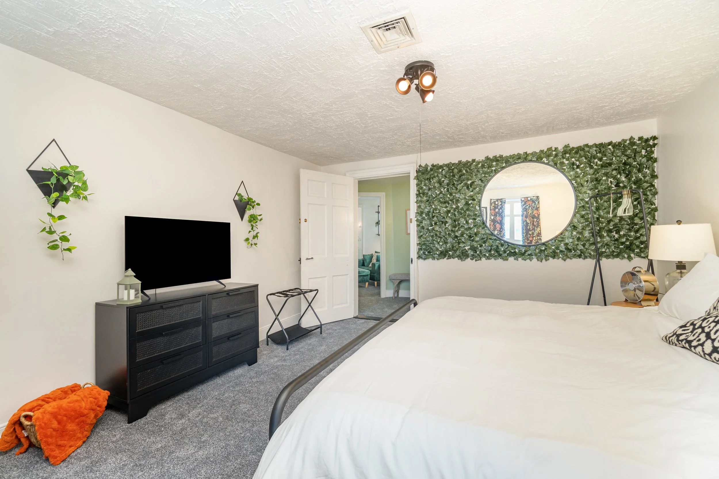 A bedroom with a white bed, wall-mounted mirror surrounded by green foliage, and a black TV stand with a television. The room has gray carpet, a textured ceiling, and a chandelier with multiple spotlights. There are decorative plants on the wall, and