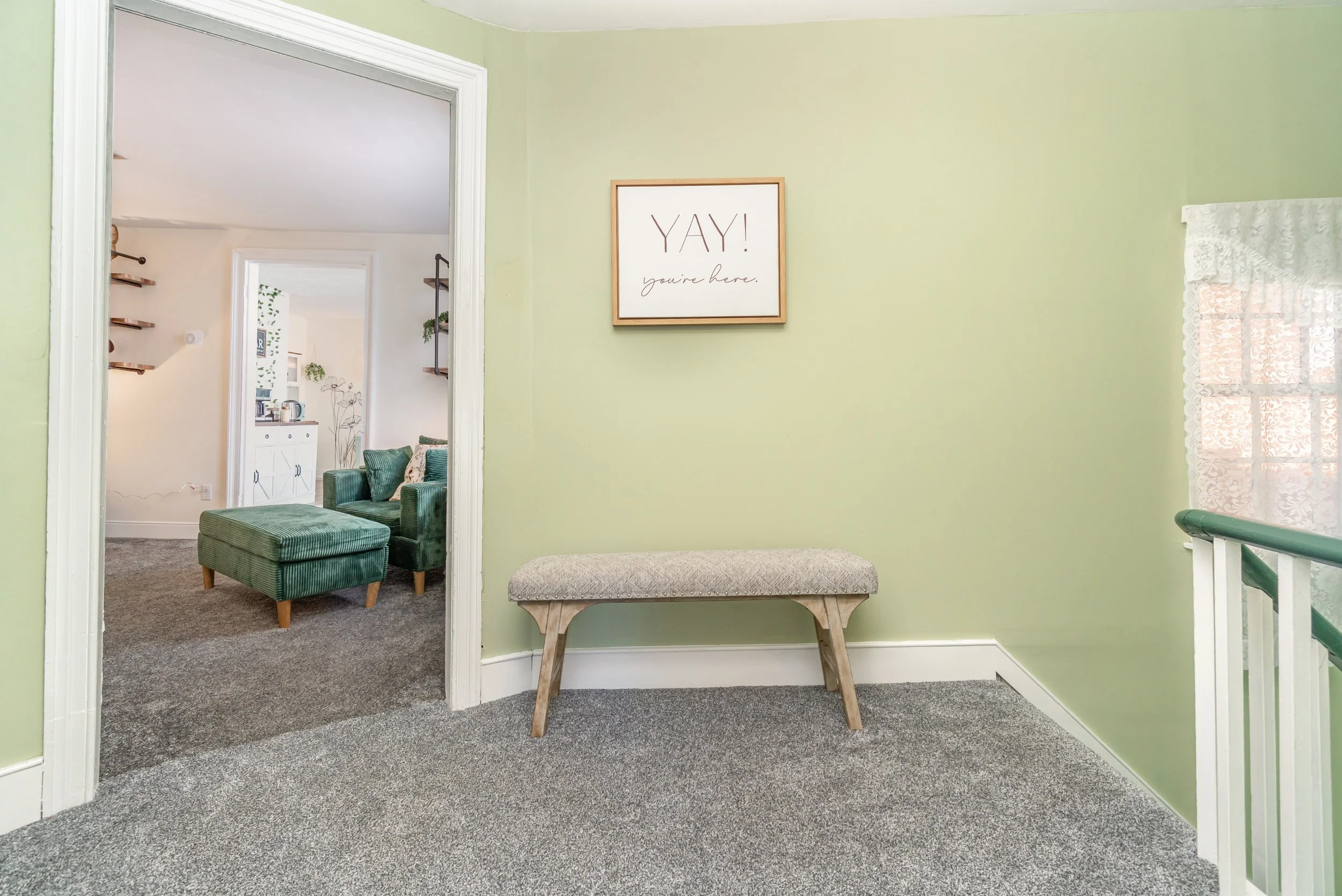 A light green wall with a framed sign that says 'YAY! you're here.' A beige bench with wooden legs is positioned against the wall. On the right, there is a window with a lace curtain, and part of a green-painted staircase railing can be seen. Beyond 
