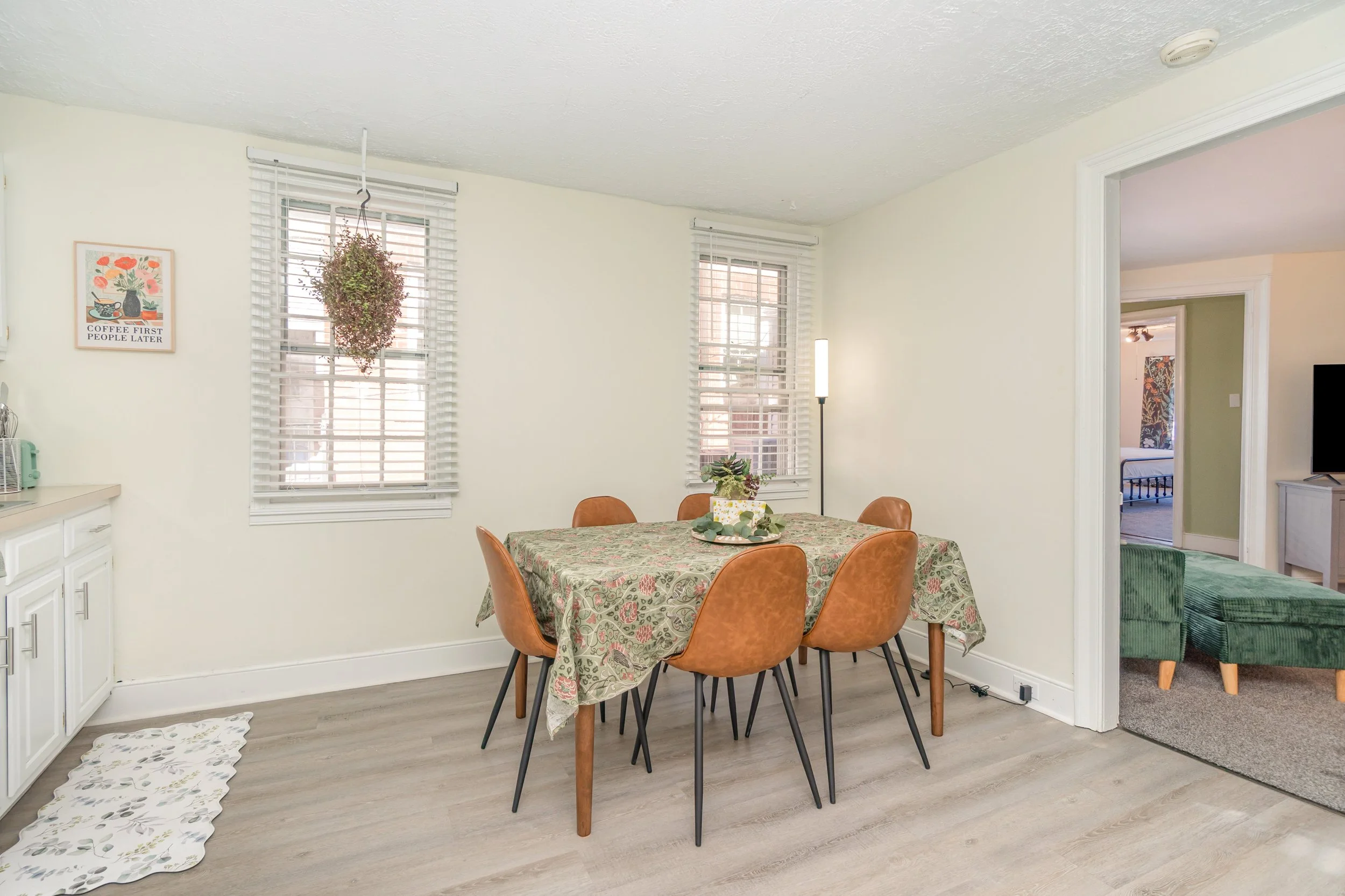 Dining room with a round table covered with a floral tablecloth, six orange chairs around it, a potted plant on the table, two windows with white blinds, a hanging plant, and a wall art with the words 'Coffee First People Later.'