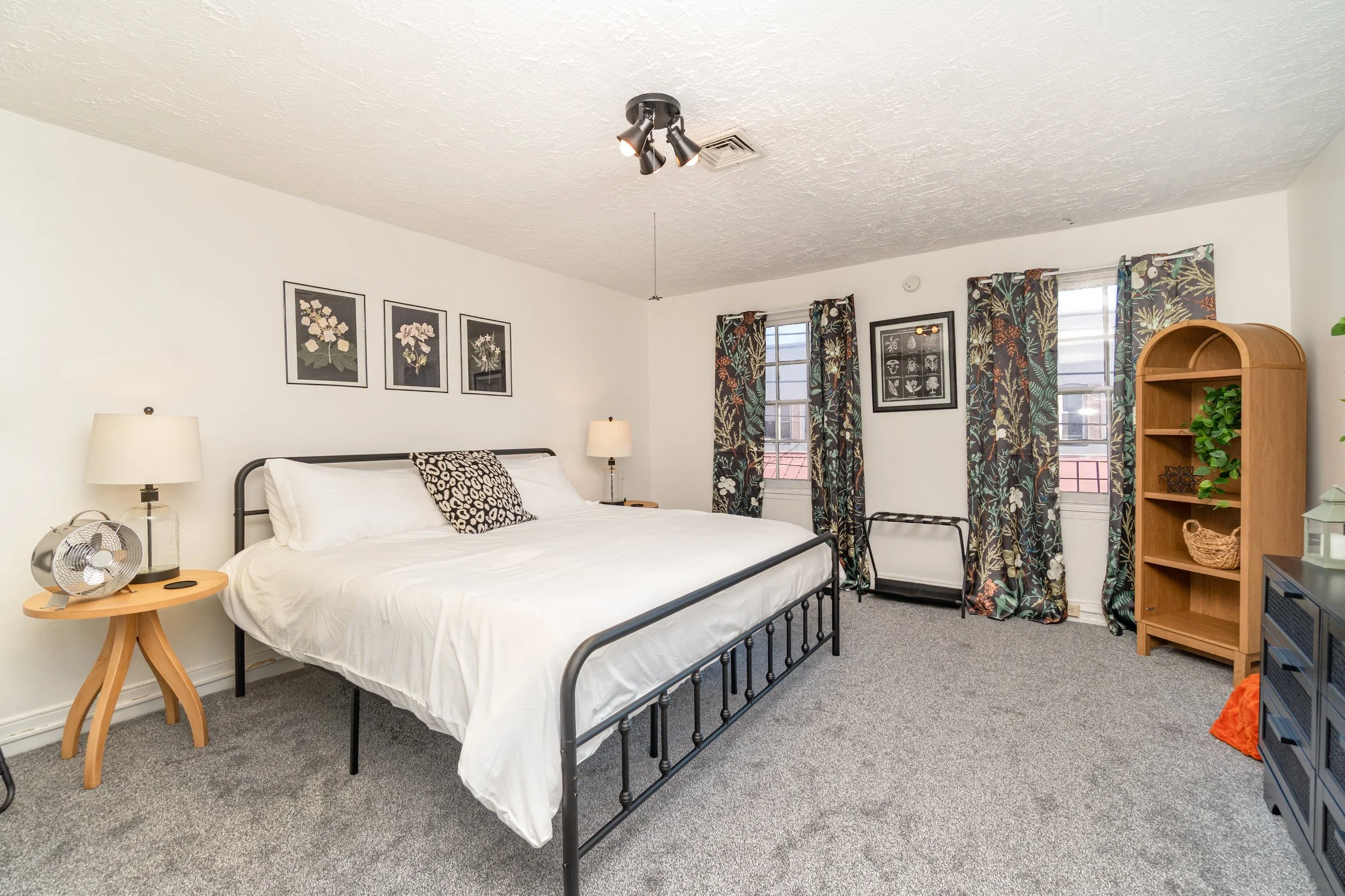 A bedroom with a black metal bed, white bedding, and a patterned pillow, two nightstands with lamps, a wooden bookshelf, and windows with floral curtains.