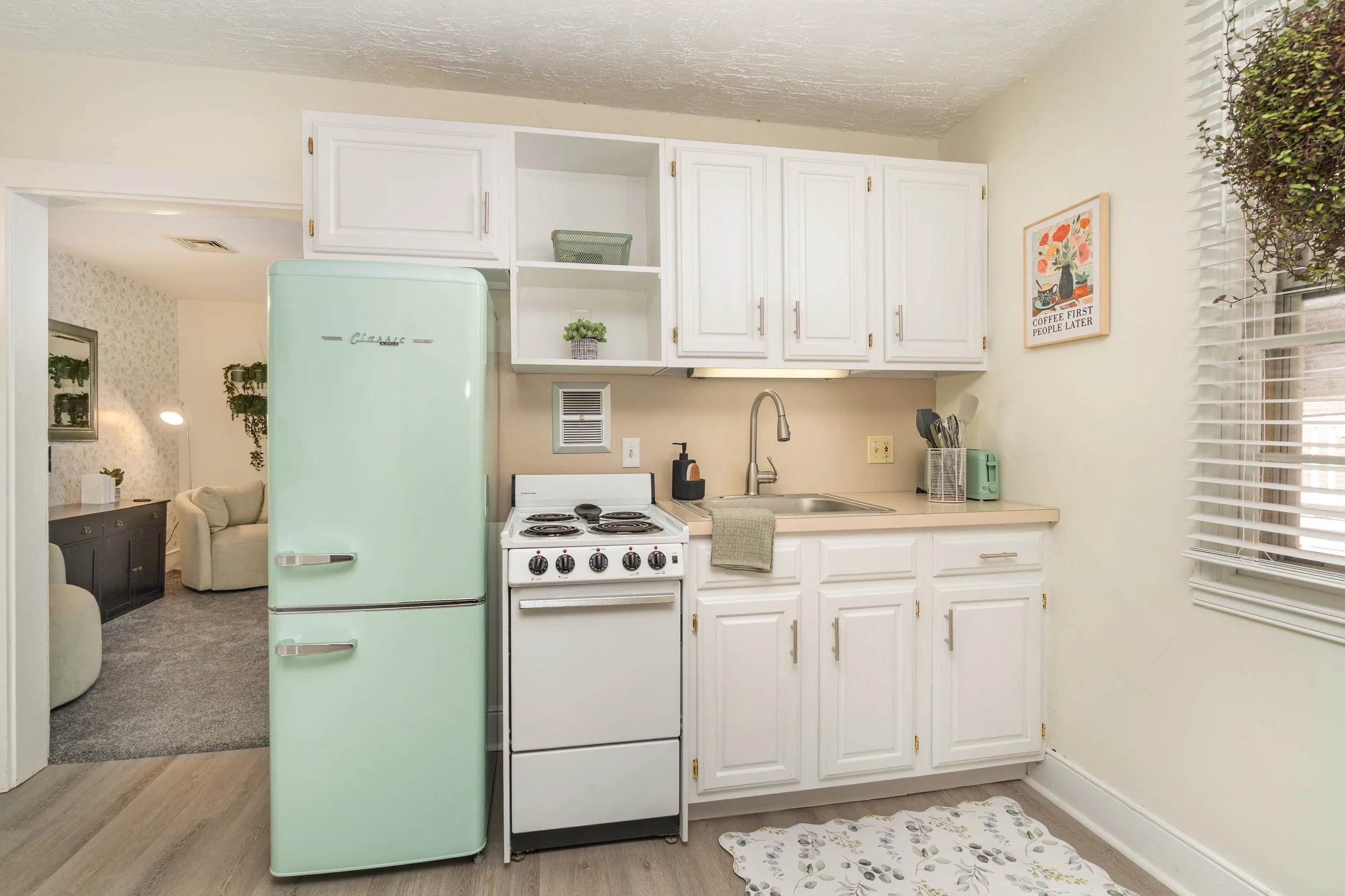 Kitchen with vintage mint green refrigerator, white stove, white cabinets, beige countertop, and a window with blinds. Decor includes a framed print on the wall, small potted plant, and kitchen utensils.