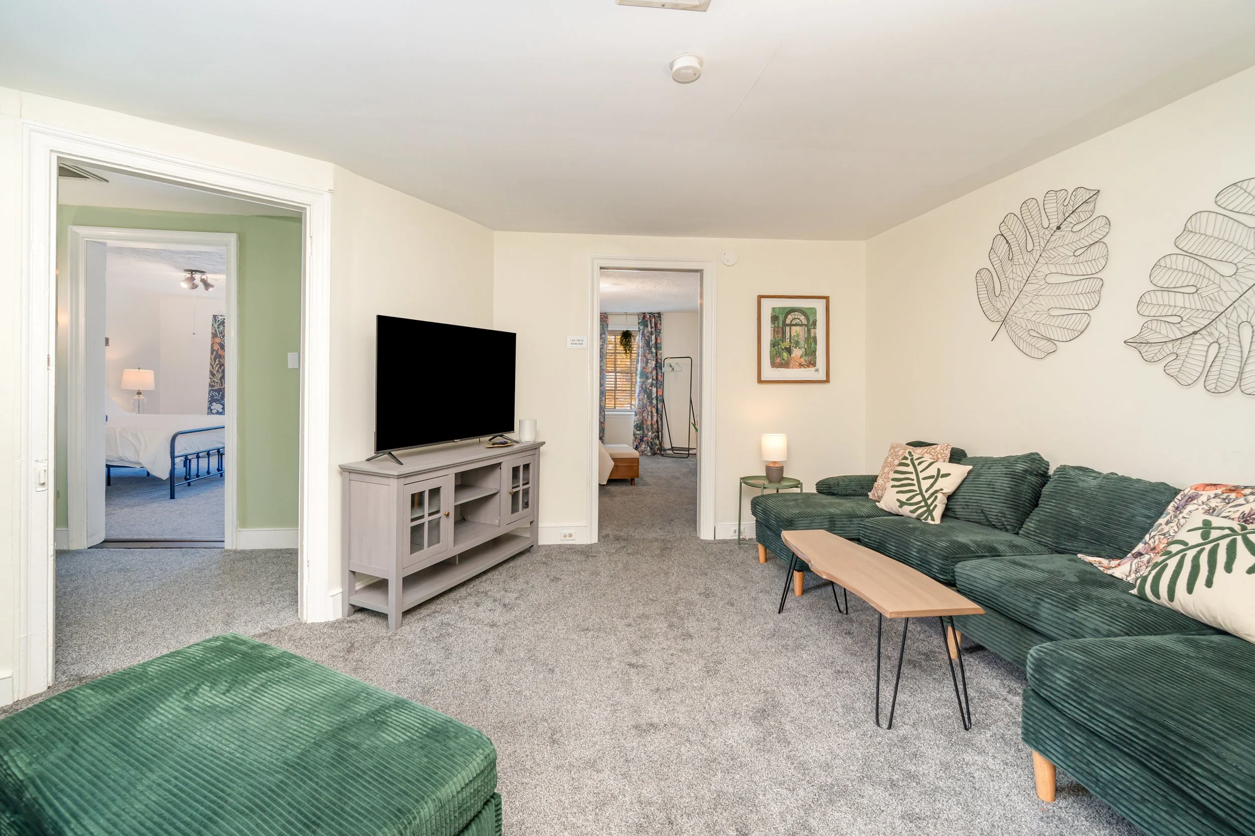 Living room with green couch, wall art of leaves, TV on gray stand, side table with lamp, doorway to bedroom with blue bed, and another doorway to a room with floral curtains.