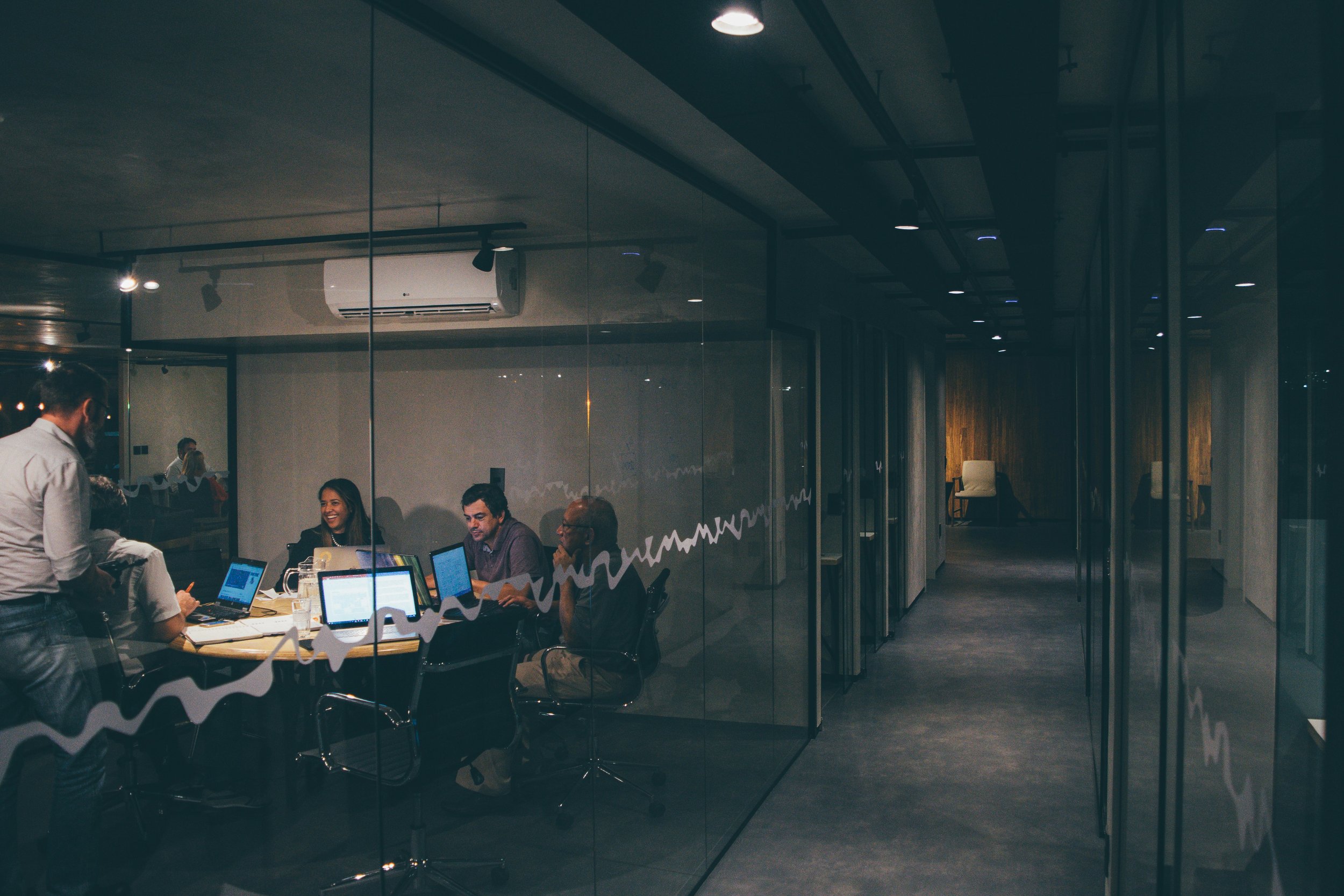 People in a modern glass-walled meeting room working on laptops and discussing, with a corridor outside the room.