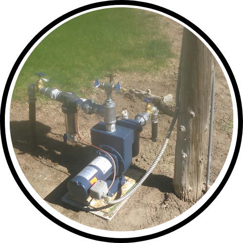 Water well pump with plumbing and valves installed on a concrete pad next to a wooden post, with grass and soil in the background.