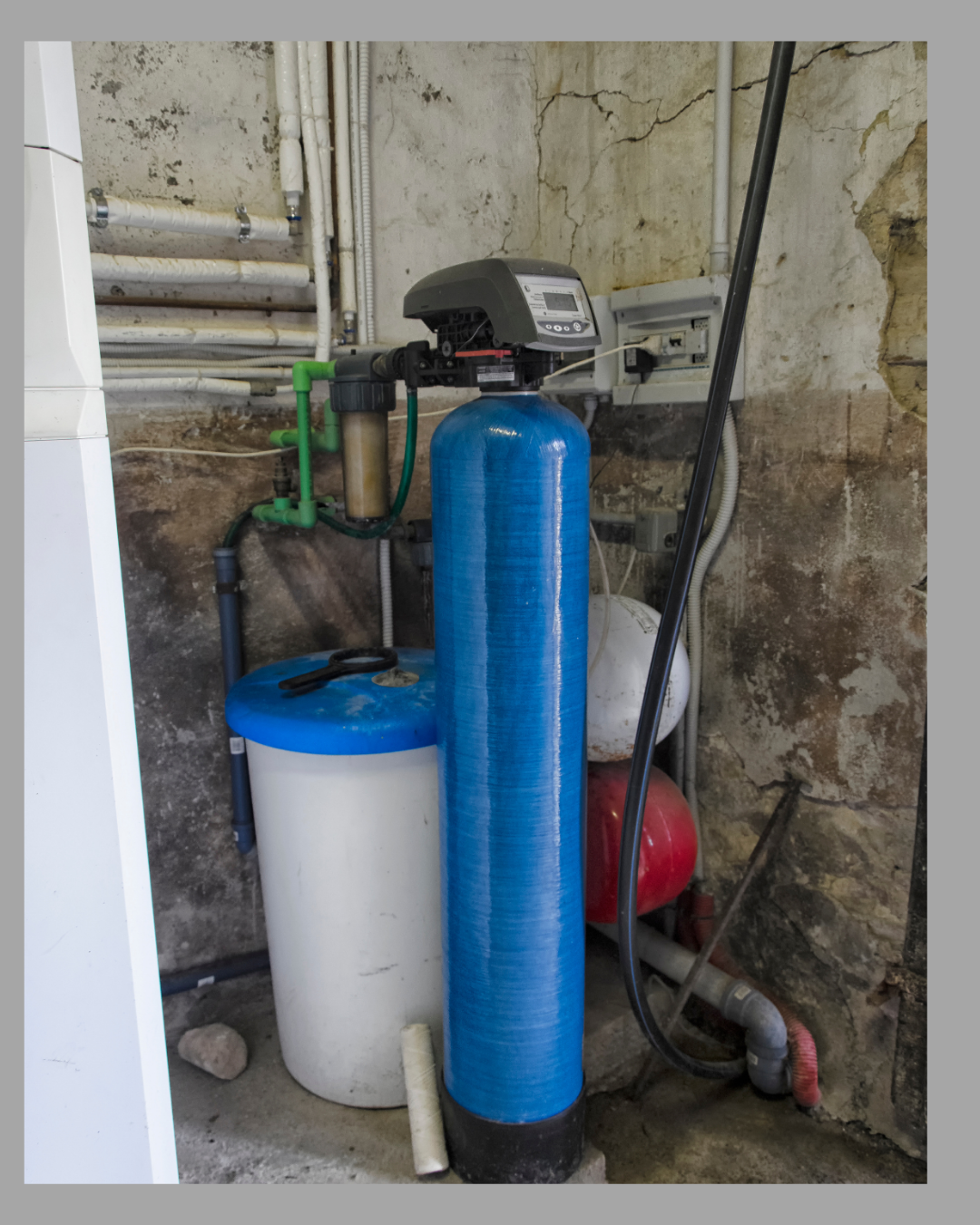 An indoor water treatment system with a large blue filtration tank, a control valve, and associated pipes and equipment installed on a rough concrete wall.