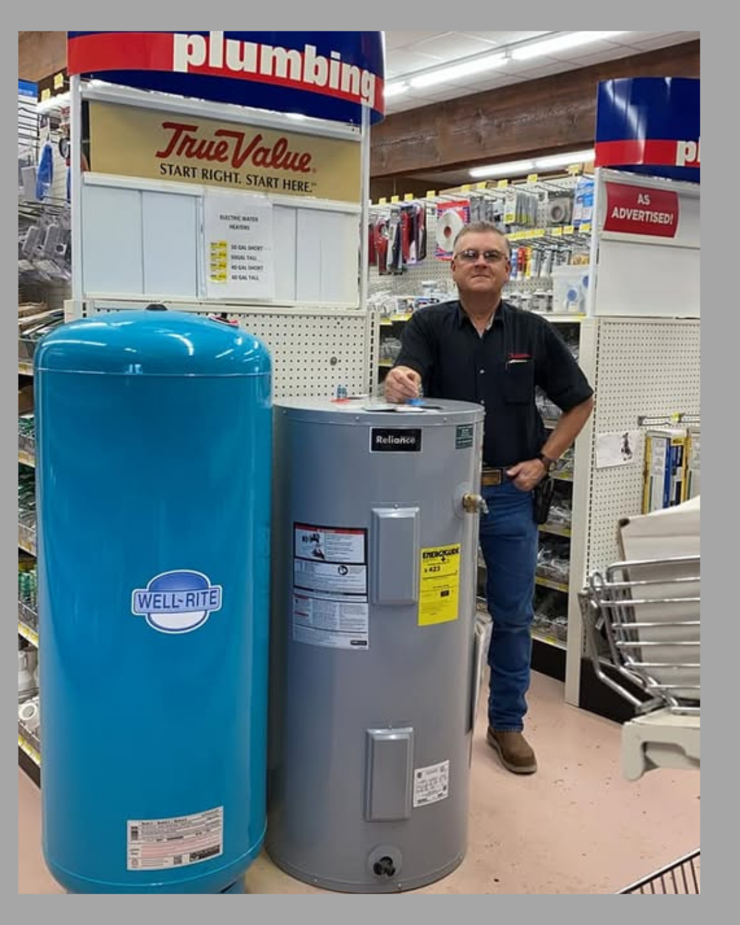 A man in a black shirt stands in a hardware store next to a large blue Well-Rite water heater and a meta Reliance water heater. The store has shelves with plumbing supplies in the background.