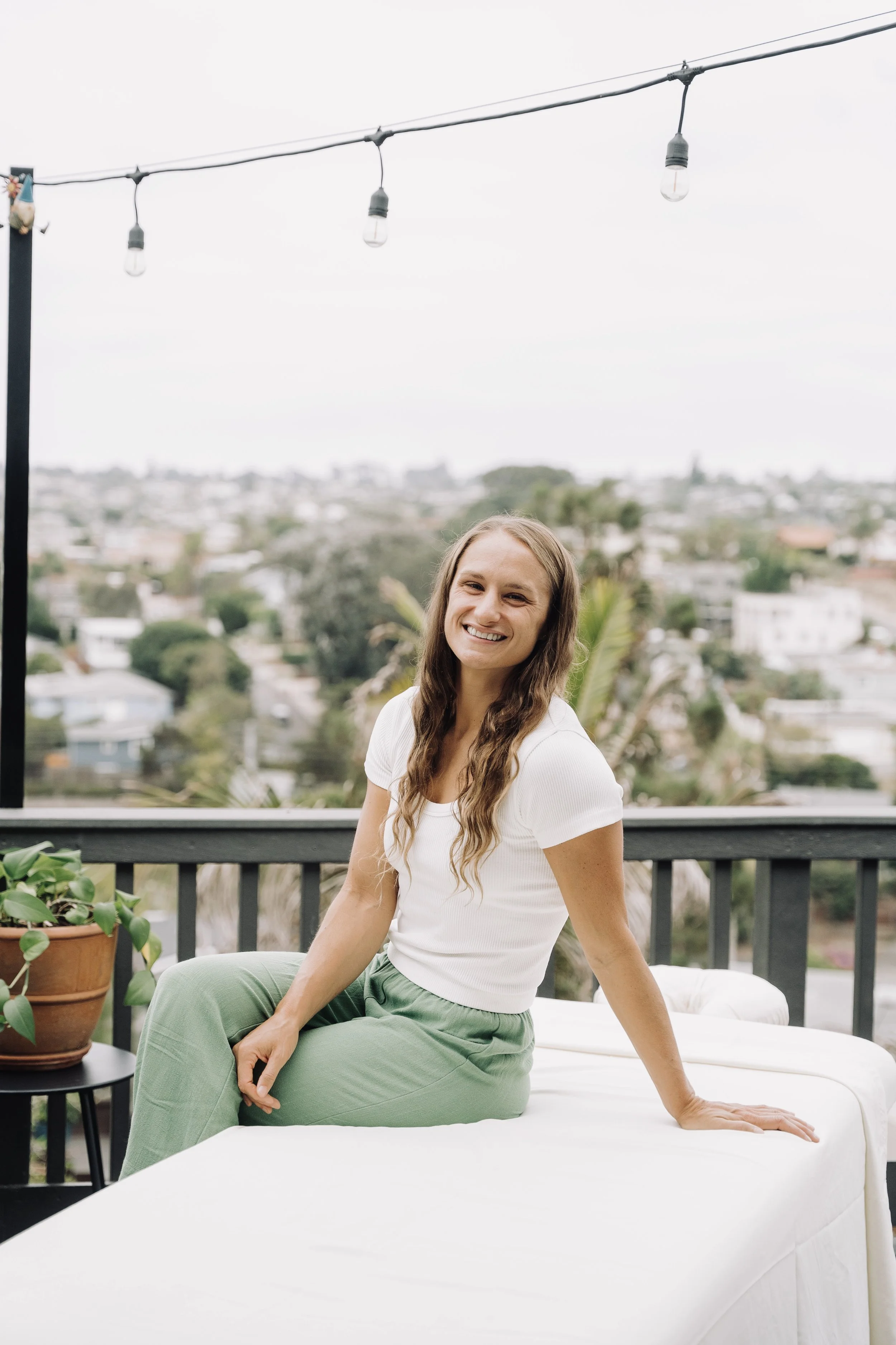 A woman with long wavy hair smiling and sitting on a white table on a balcony with a city view and outdoor string lights overhead.