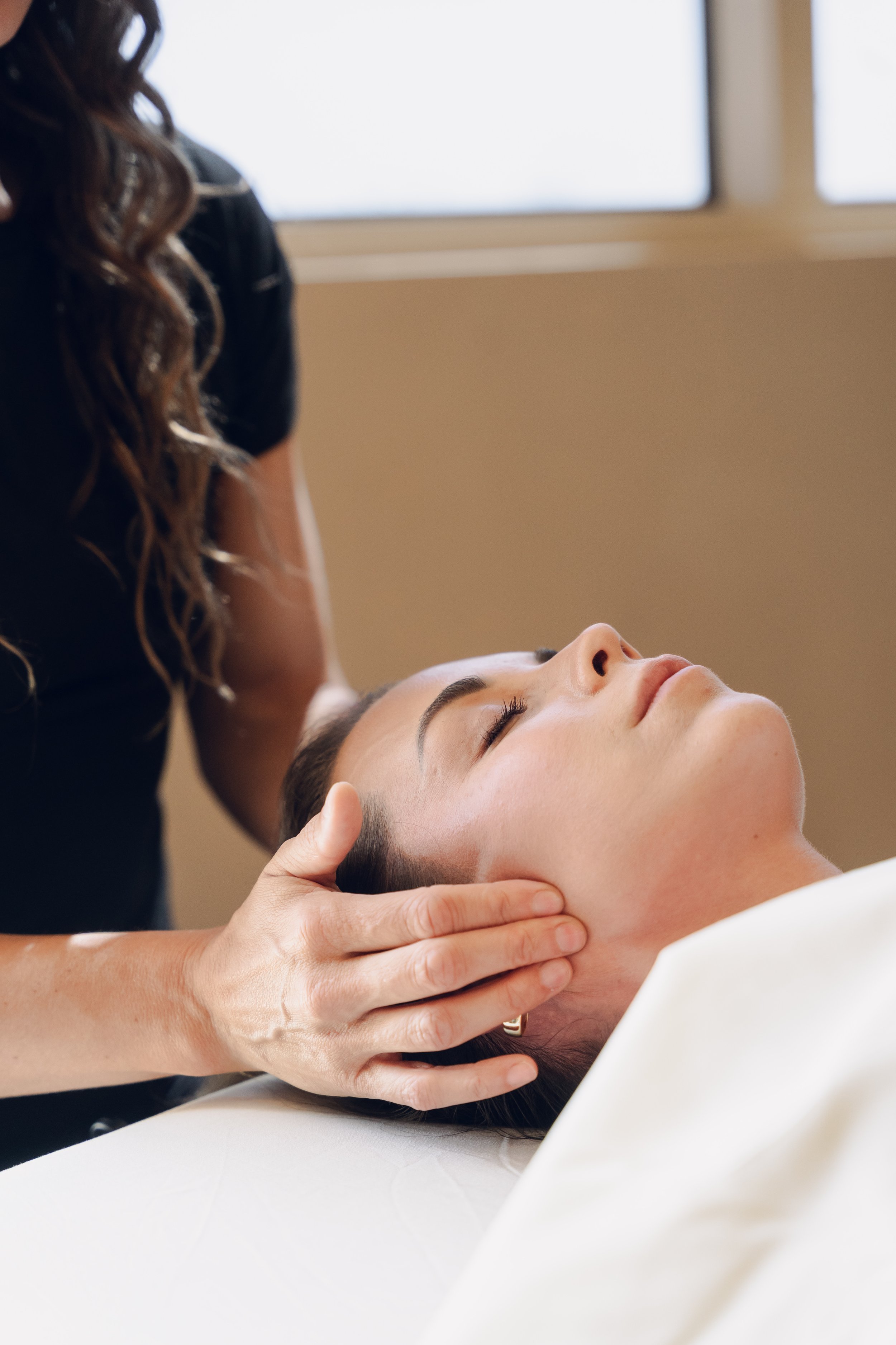 A woman receiving a massage or facial treatment, lying on a table with her eyes closed, as a therapist gently holds her head and neck.